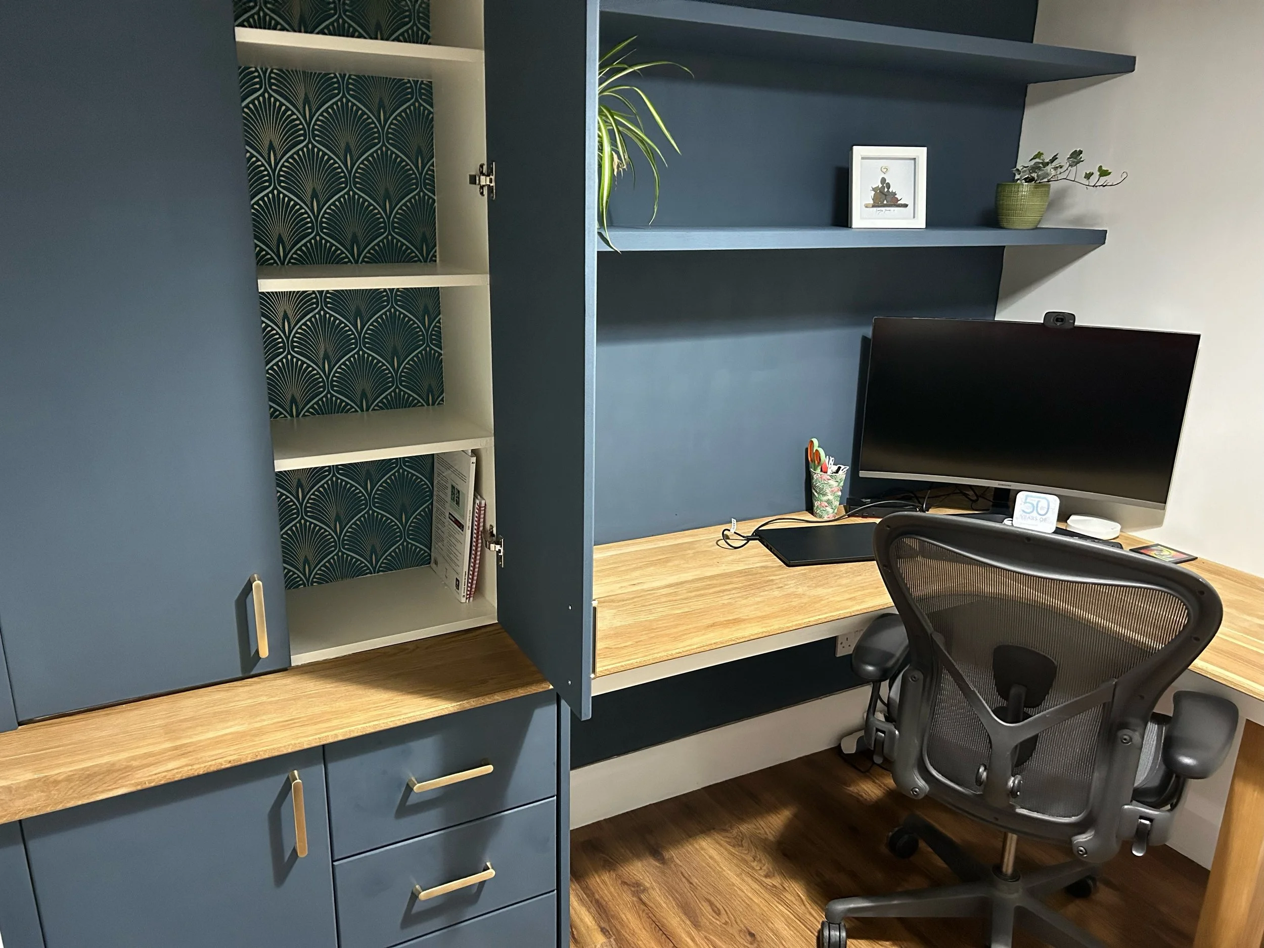 Home office desk with computer monitor, office chair, shelves with picture frame and plant, and blue cabinetry.