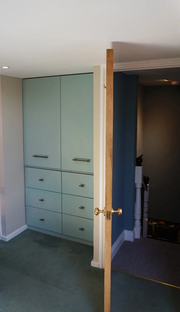 Partially open door in a home, revealing a built-in cabinet with light blue doors and drawers, next to a staircase with a white balustrade, in a room with a gray carpet and white walls.