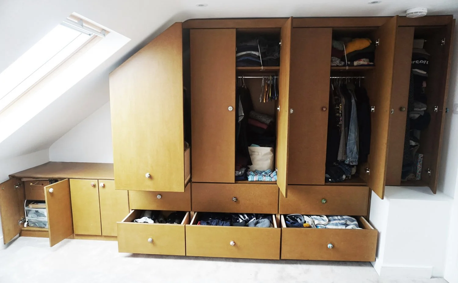Wooden built-in wardrobe with open doors revealing clothes, shoes, and storage compartments, situated underneath a sloped ceiling with a skylight.