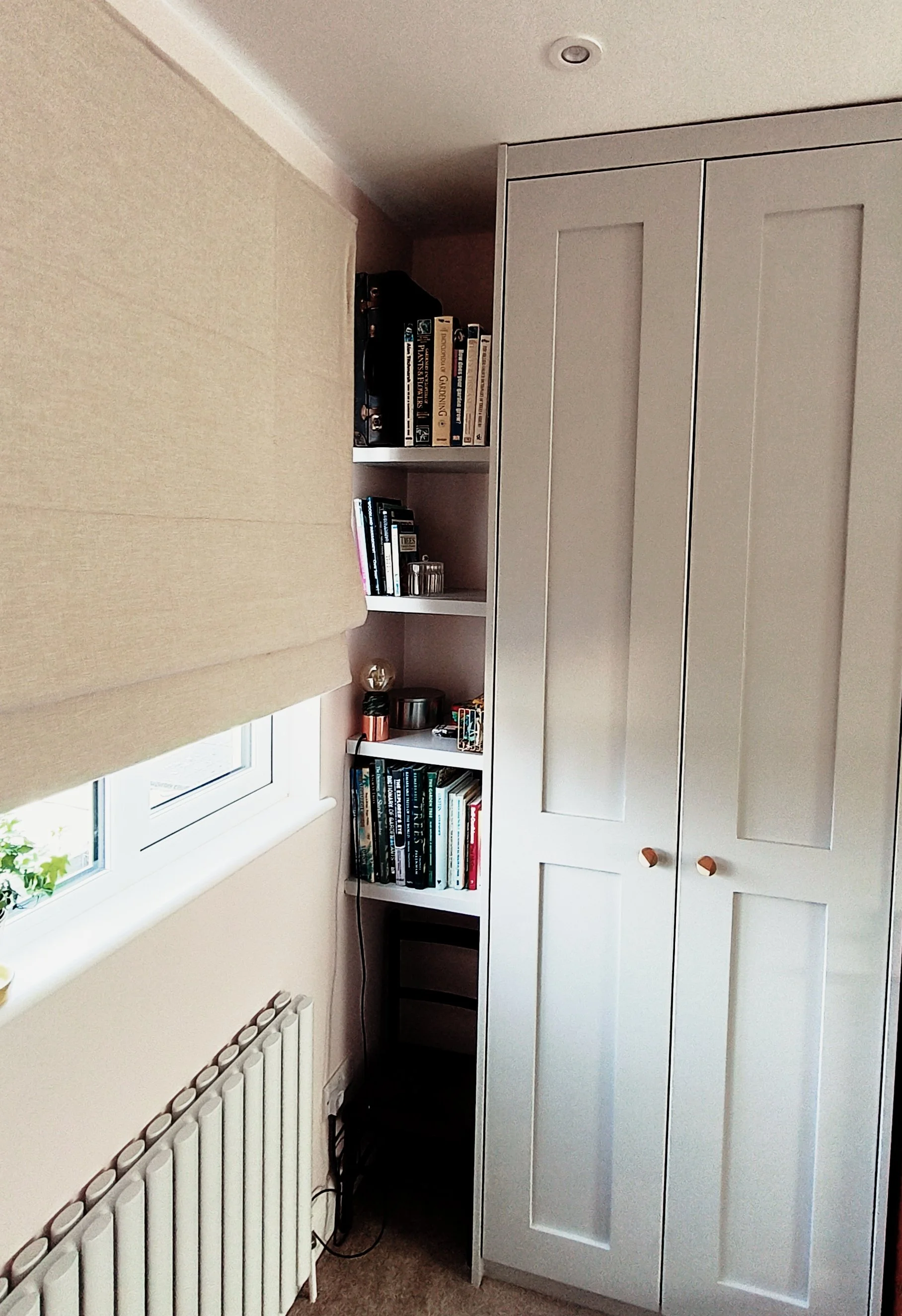 A corner of a room with a white window, beige curtains, a white bookshelf filled with books, a small black table, and a tall white wardrobe with wooden handles.