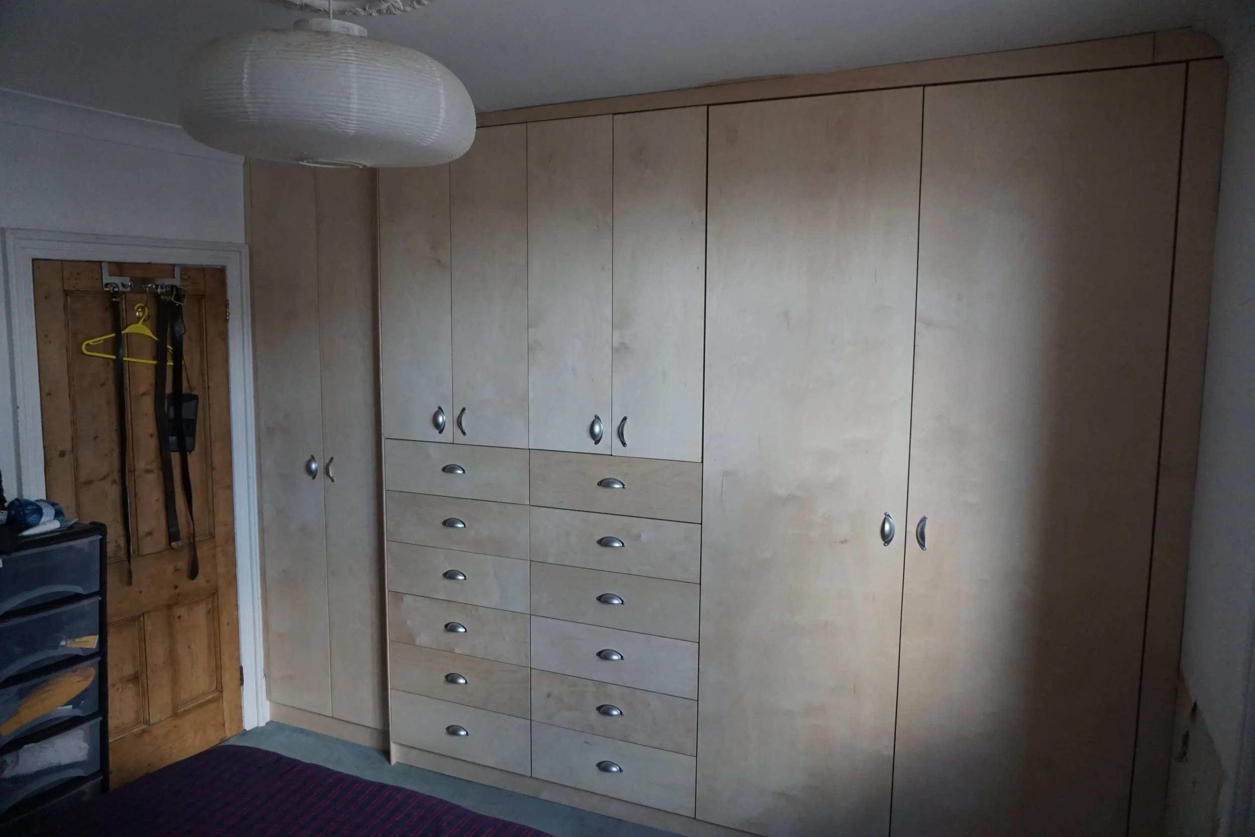 Bedroom with large custom wood wardrobe with multiple doors and drawers, a wooden door with hooks and hanging items, and a plastic storage unit, with a ceiling-mounted paper lantern light.