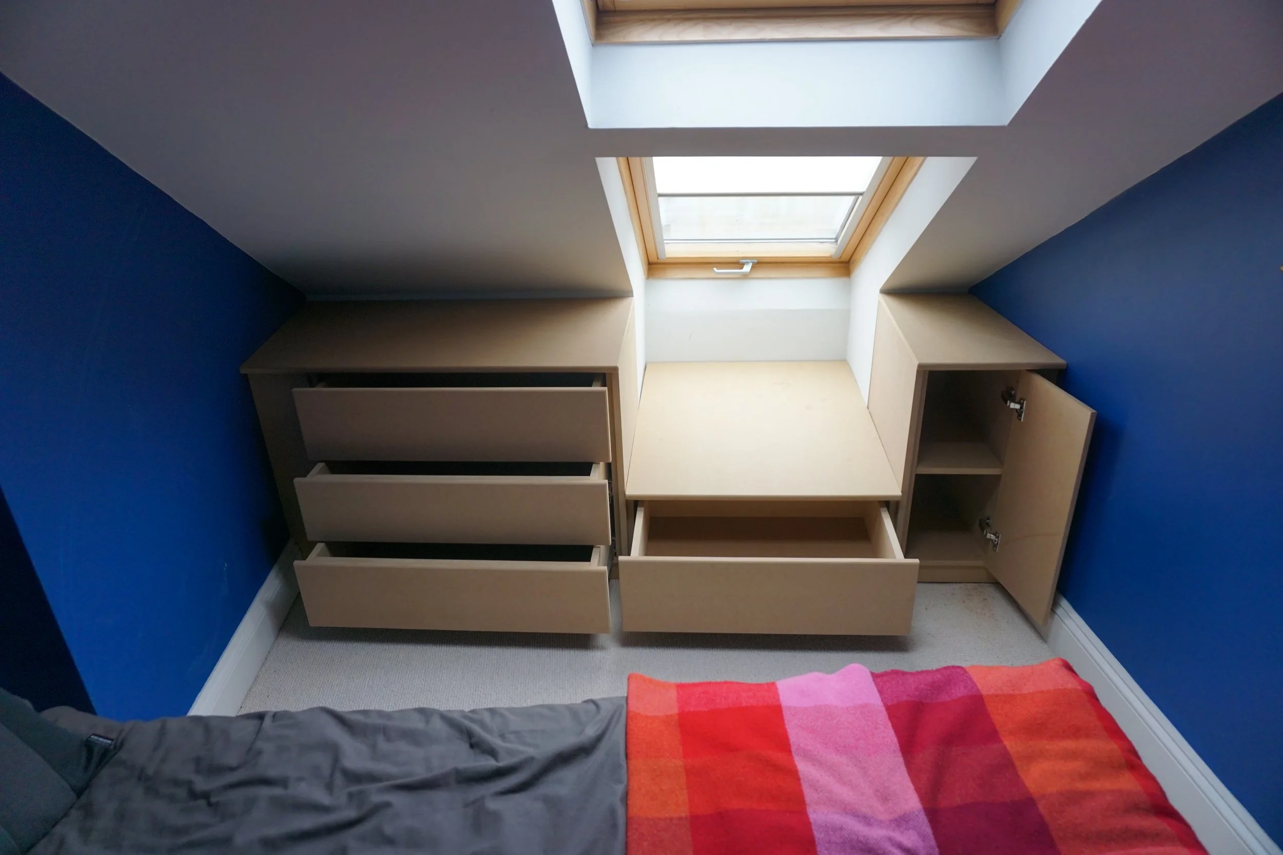 A room with blue walls and a sloped ceiling featuring two skylights. There are beige built-in cabinets below the skylights, with drawers and open shelves. A part of a bed with a gray blanket and a red, pink, and orange checkered blanket is visible in