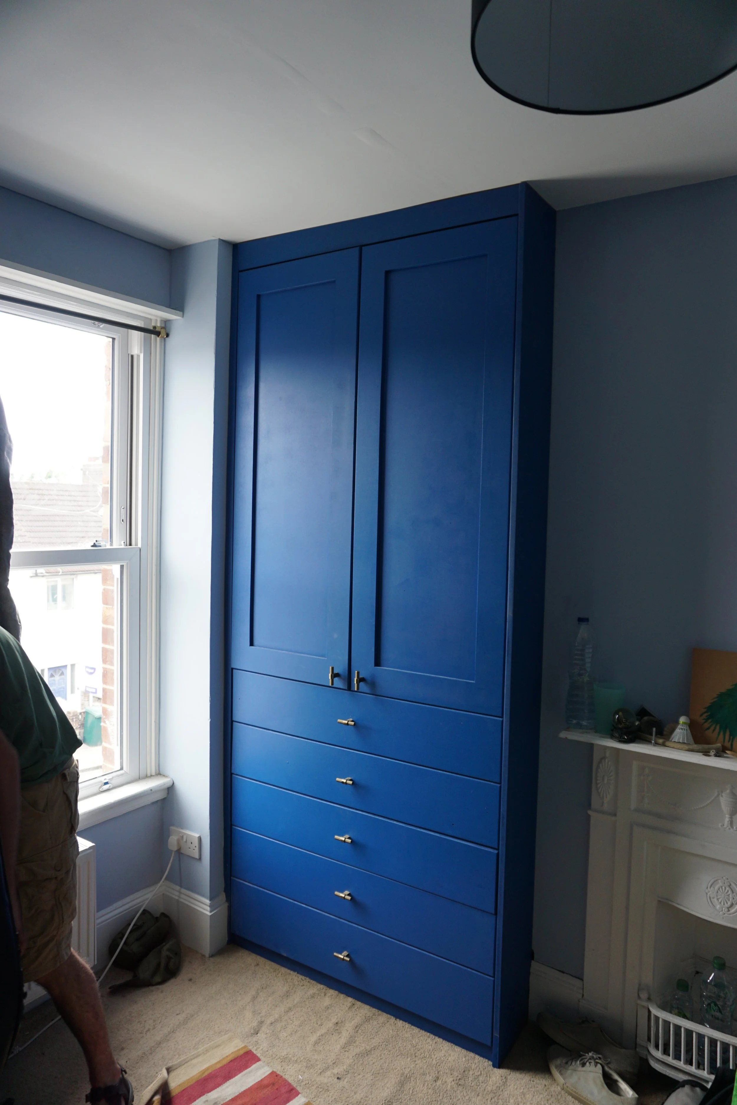 A tall blue wardrobe with drawers on the bottom right corner in a room with a window and a white fireplace.