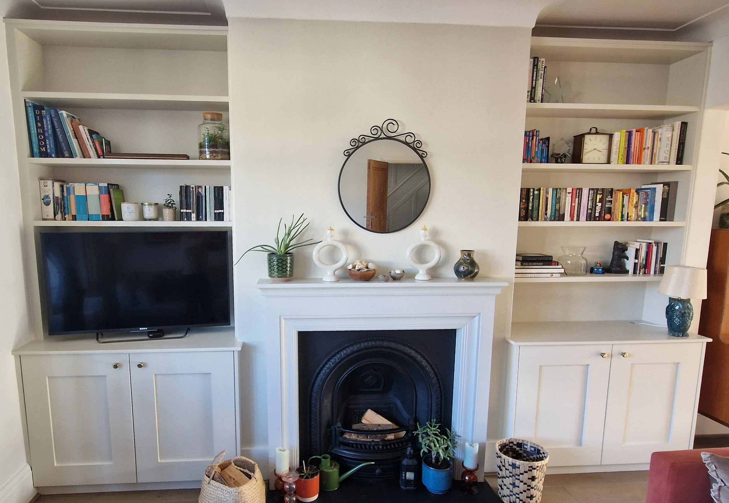 Living room with white built-in shelves on either side of a black fireplace, decorated with books, plants, and decorative objects, with a small mirror hanging above the fireplace.