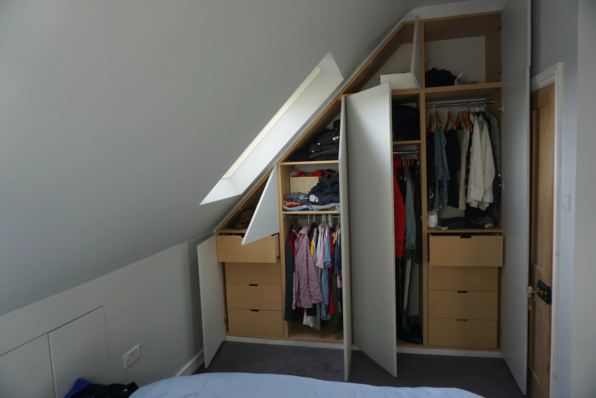 Built-in wardrobe with open doors revealing clothes and storage compartments under a sloped ceiling.