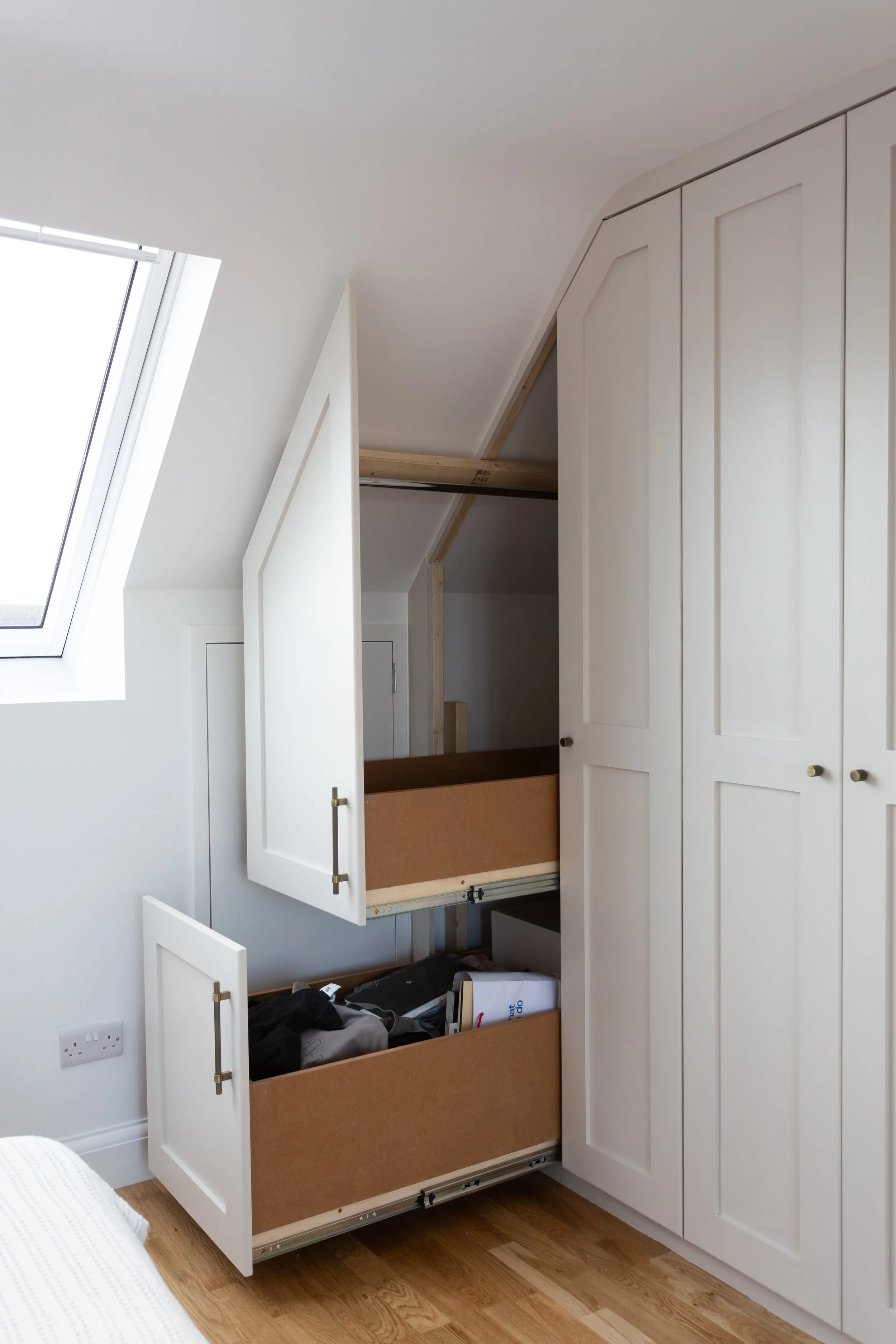 Built-in white wardrobe with open sliding drawers and cabinets in a bedroom under a sloped ceiling with a skylight window.