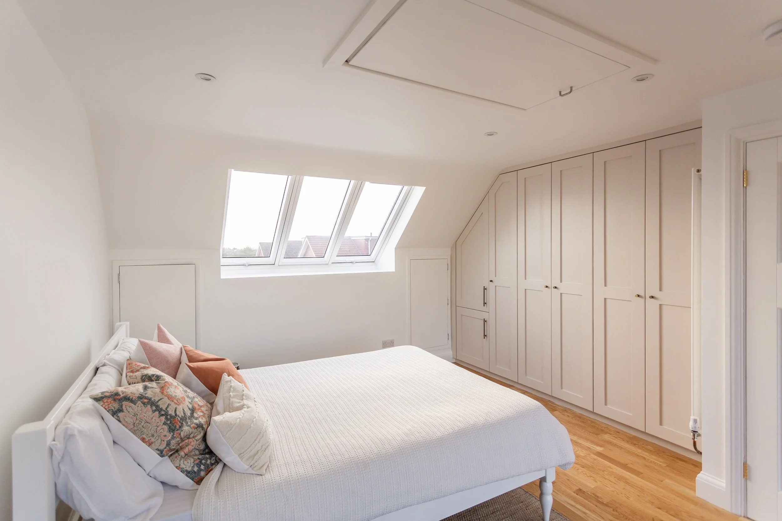 A bedroom with white walls and ceiling, a bed with white bedding and decorative pillows, hardwood floors, and a built-in wardrobe. There are three skylight windows on a sloped ceiling.