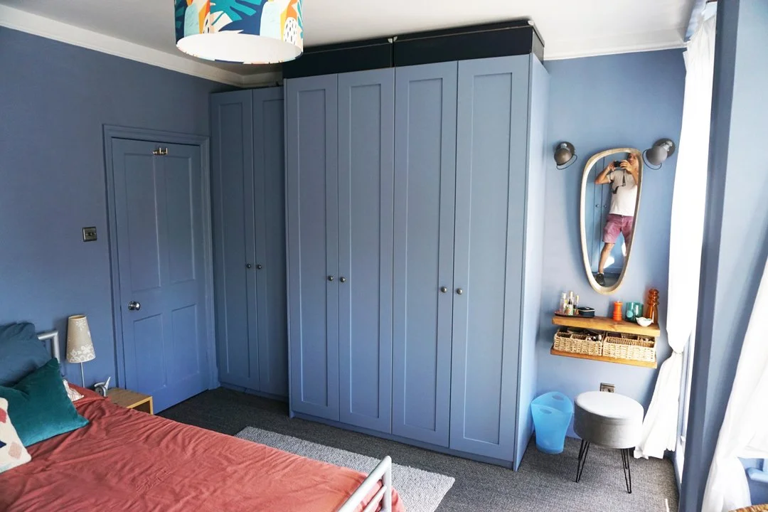 A bedroom with blue walls and large blue wardrobe, a bed with a pink bedspread, a small side table with a lamp, a mirror with a person taking a photo, a wicker shelf with personal items, and white curtains.