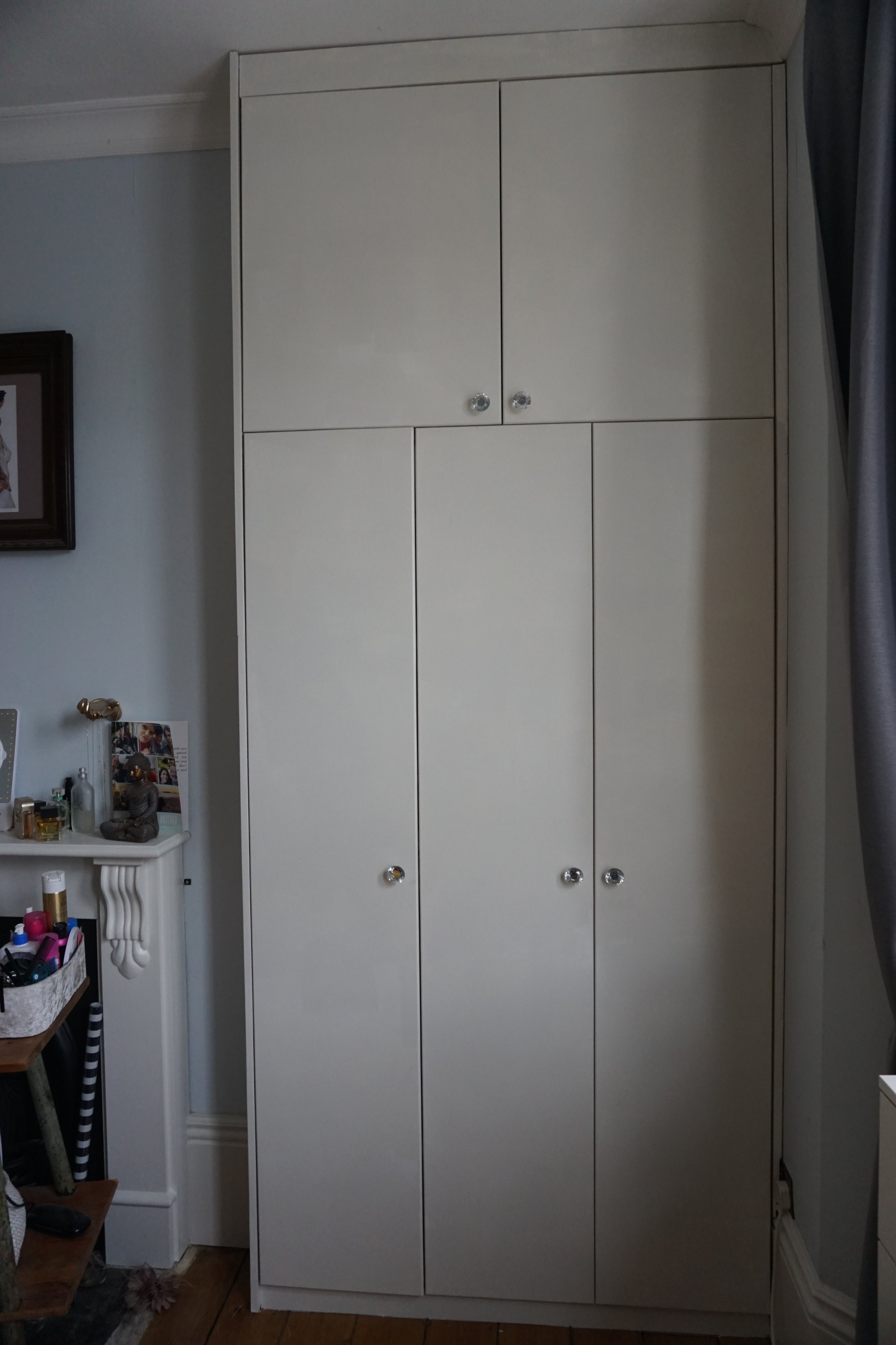 A tall, white storage cabinet with two large doors on the lower section and two smaller doors on the upper section, with clear round knobs, located in a room with light blue walls and hardwood flooring.