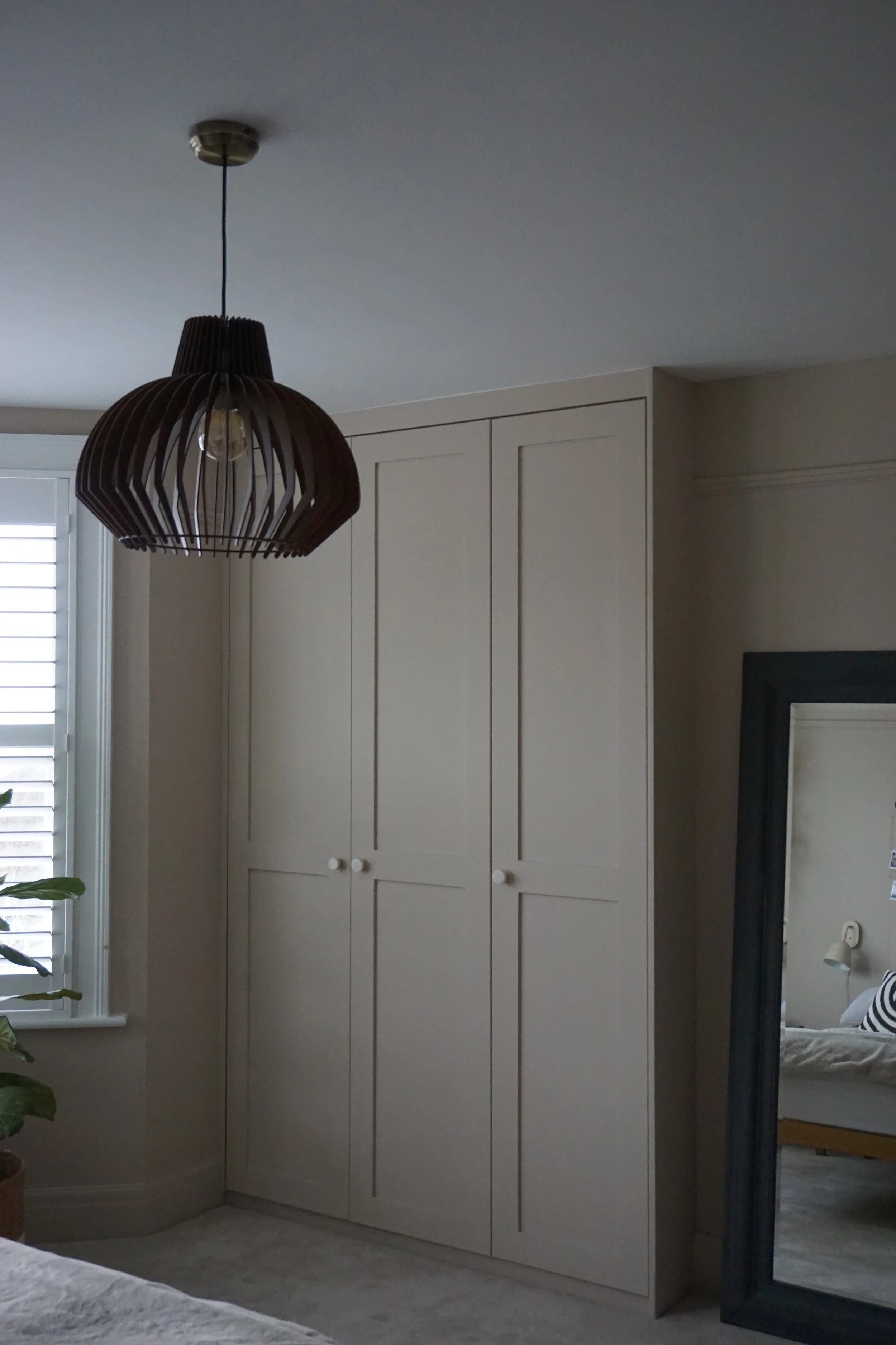 A corner of a bedroom with a large white closet, a black full-length mirror, a window with blinds, and a hanging wooden pendant light.