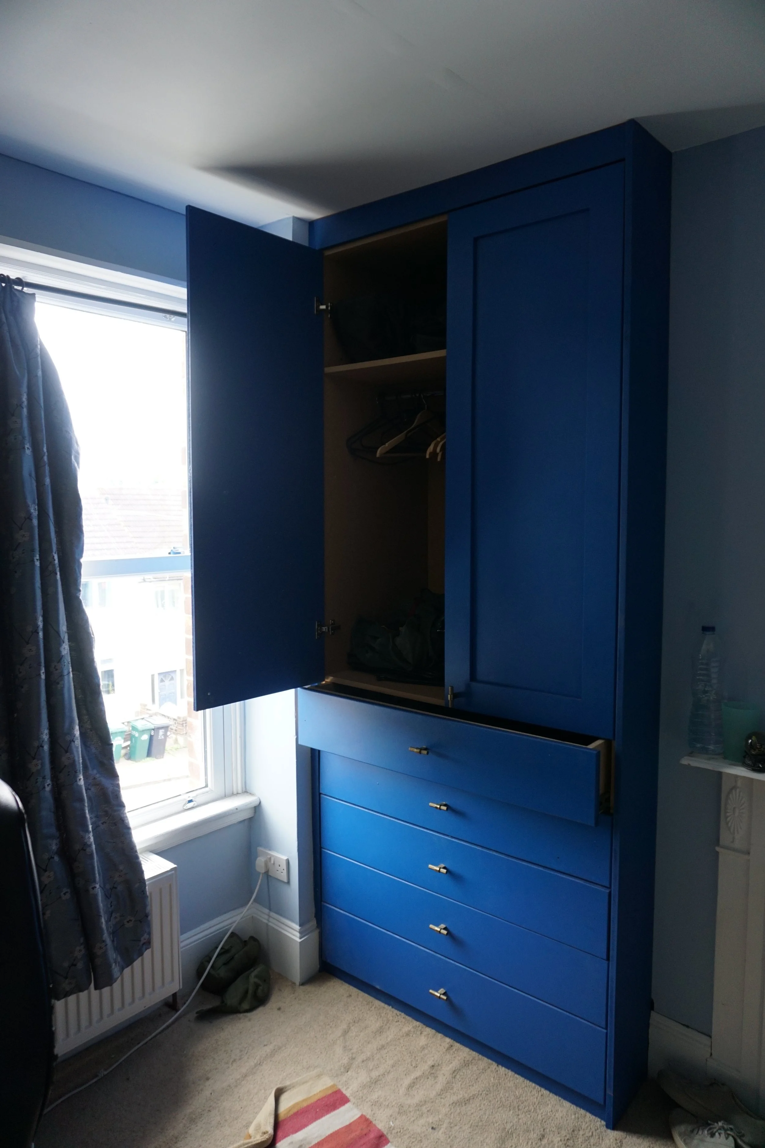 Open blue wardrobe with shelves, drawers, and hanging space in a bedroom, near a window with curtains.