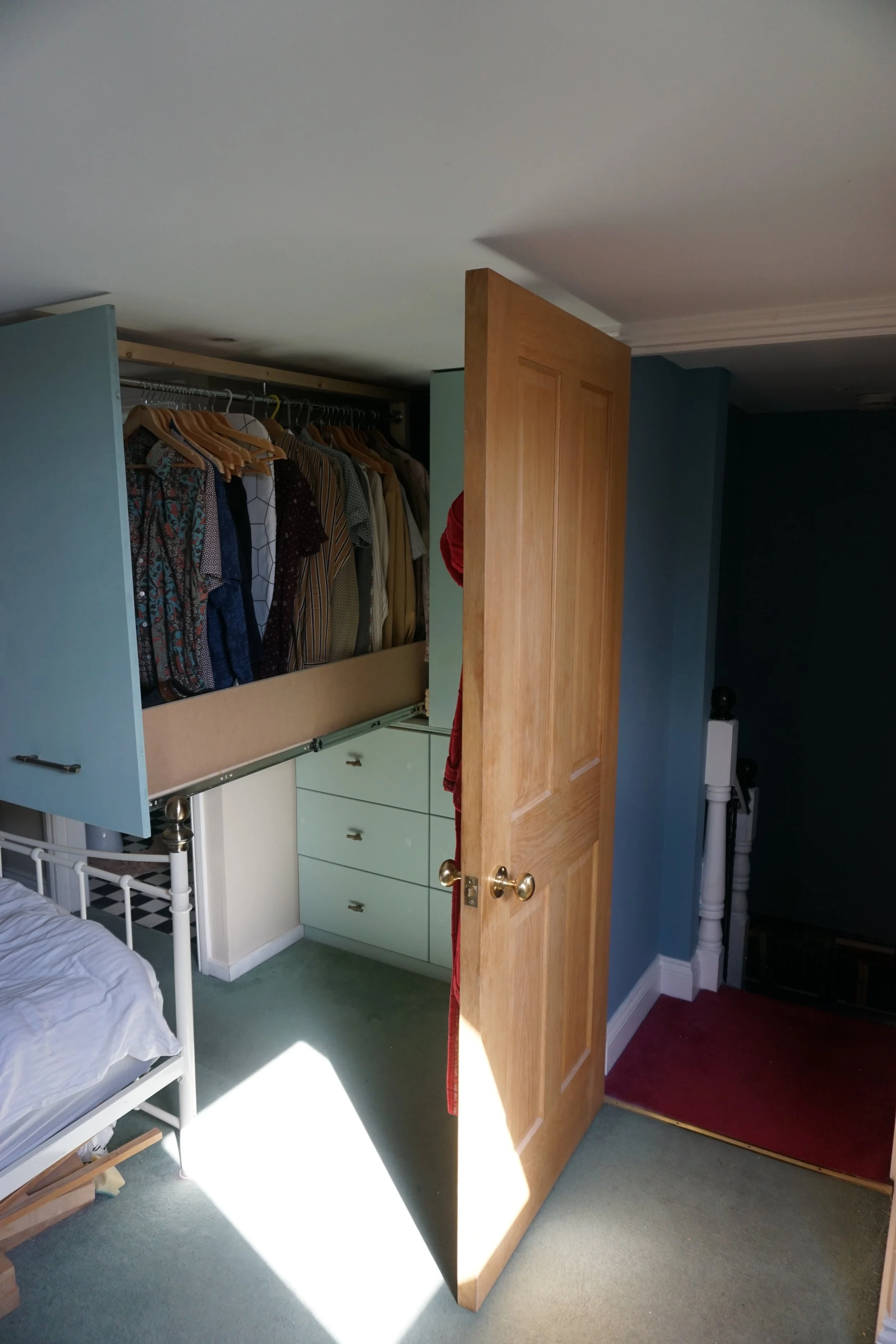 Open closet door revealing a hanging rack with various patterned shirts, a light green dresser drawers, part of a white bed frame, and a stairway with a red carpet.