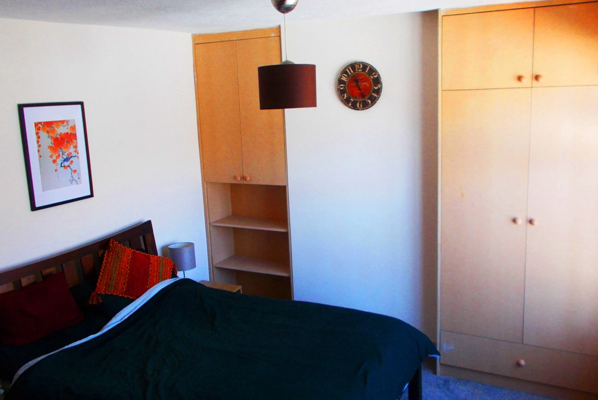 A bedroom with a bed, a framed picture of leaves and a bird on the wall, a nightstand with a lamp, a black and red wall clock, a hanging pendant light, and built-in wooden wardrobes.