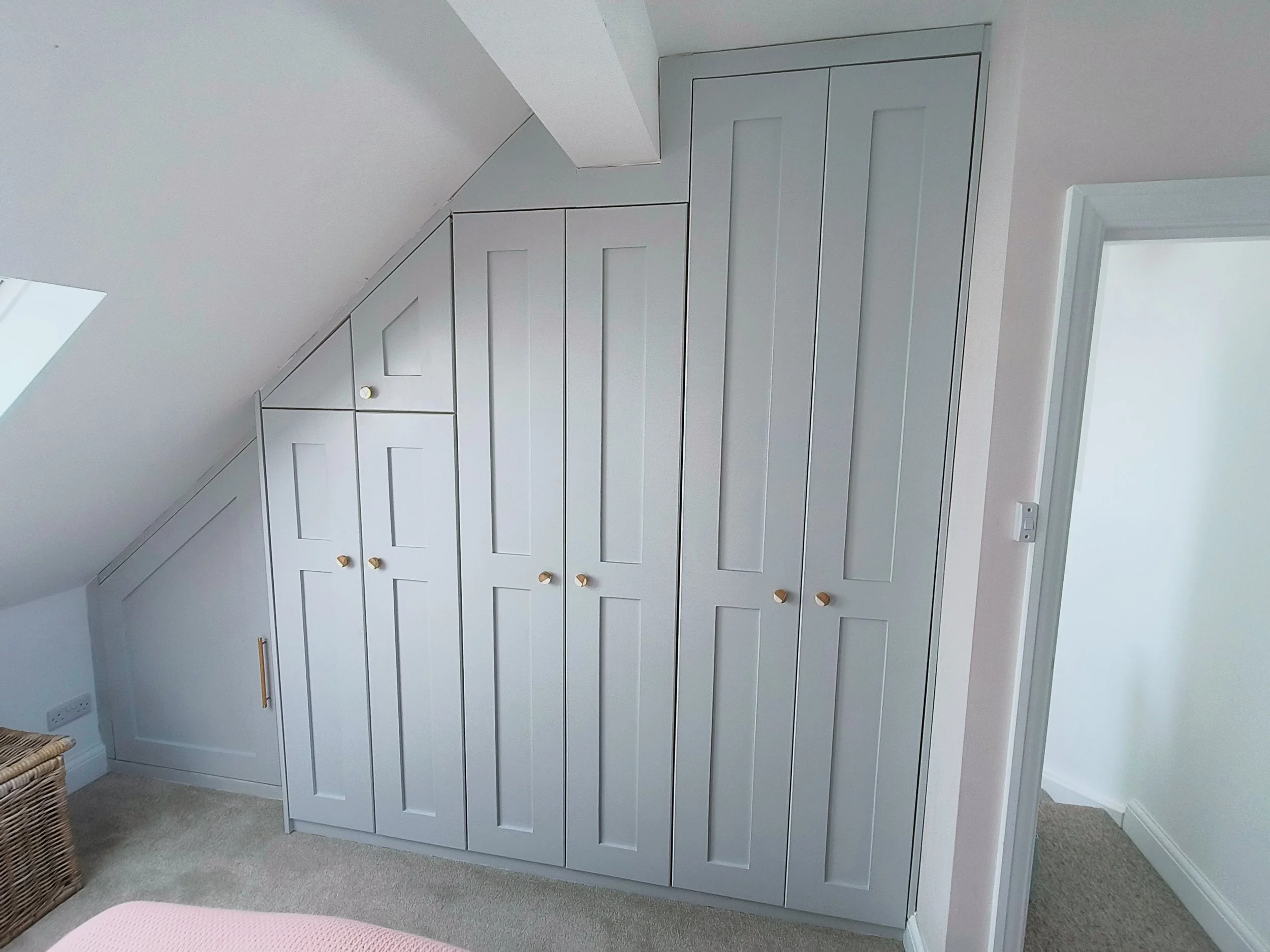 Light gray built-in wardrobe with multiple doors and wooden knobs in a room with beige carpet and white walls.