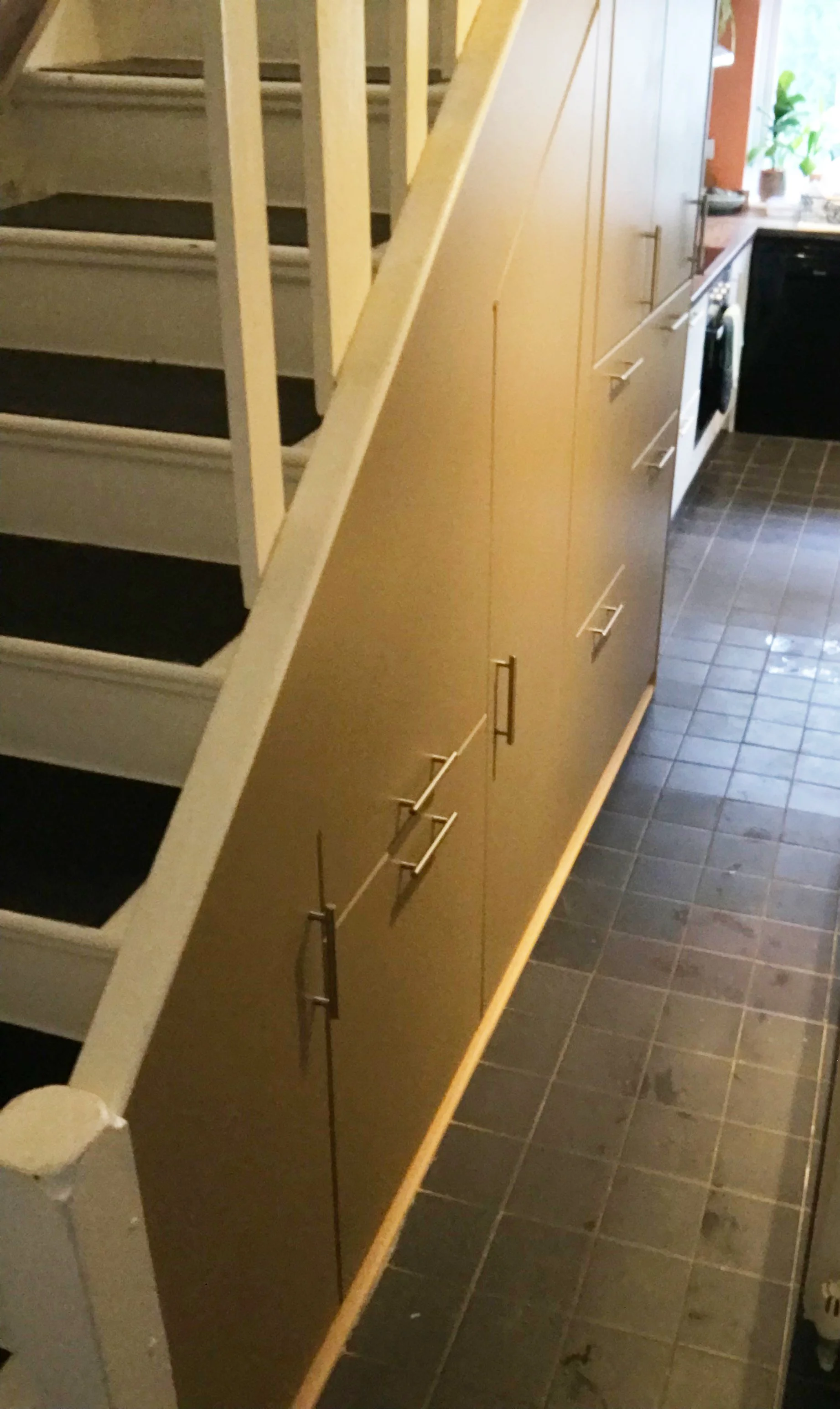 View of a staircase with black steps and white railing next to a beige cabinet unit with silver handles in a kitchen with dark tiled flooring.