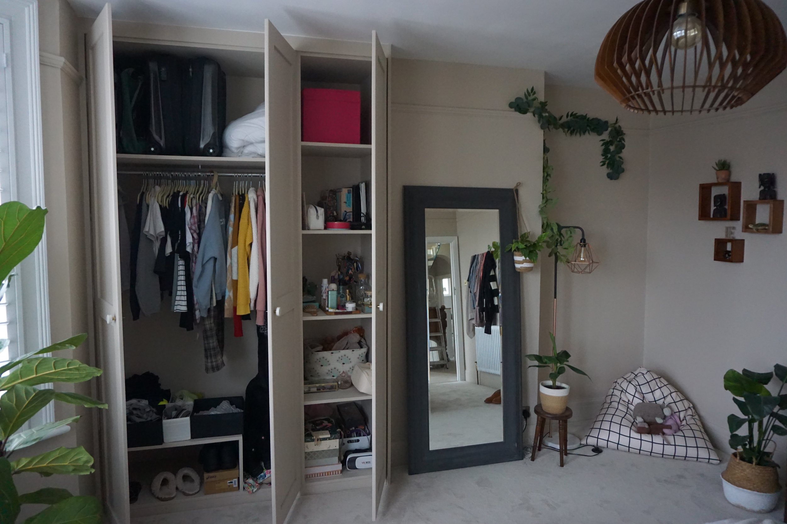 Bedroom with an open wardrobe, a large framed mirror, a potted plant, a reading nook with a checkered cushion, and wall decor.