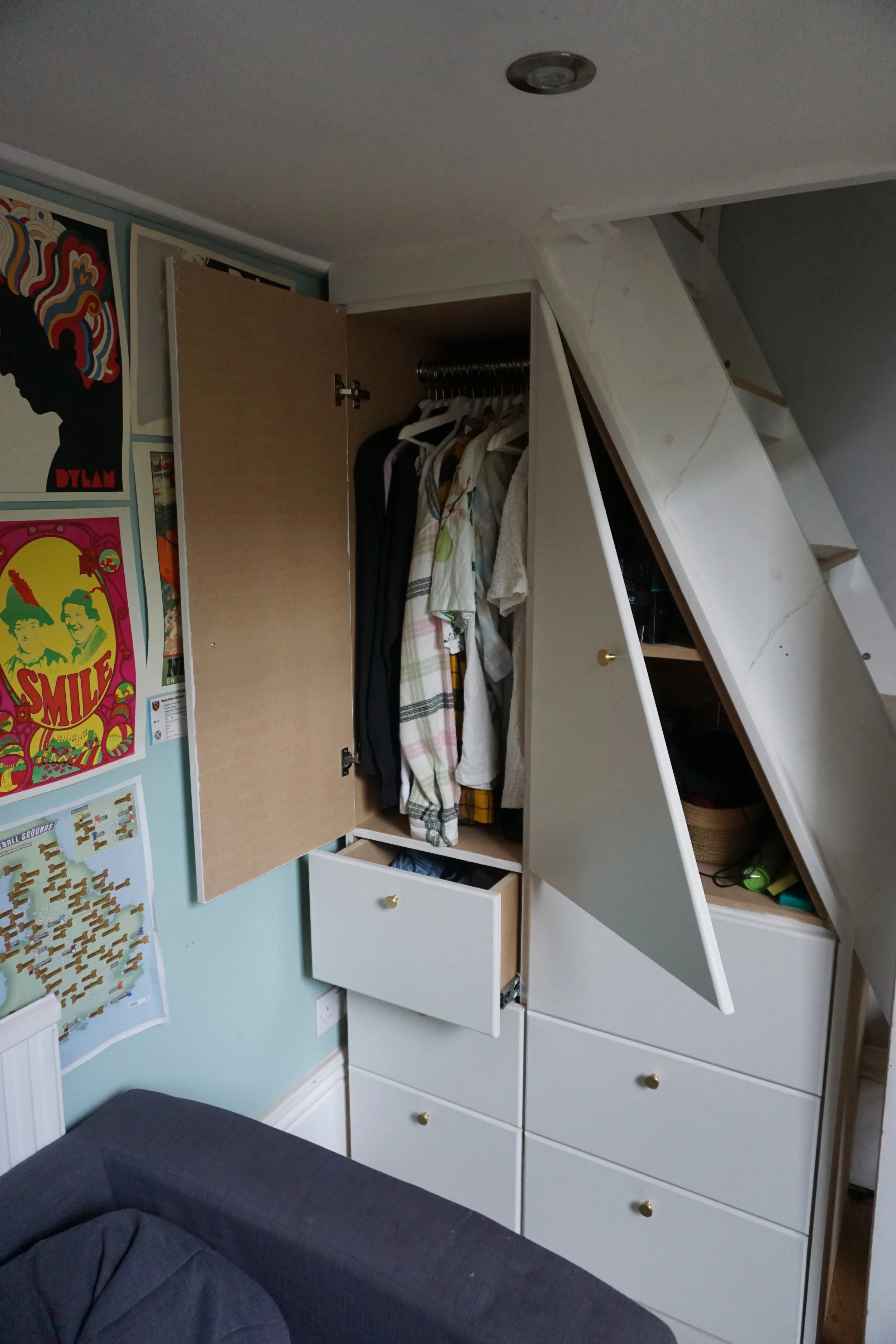 Built-in closet with open doors revealing hanging clothes and drawers underneath, next to a wall with posters and a map, and a corner of a gray sofa in the foreground.