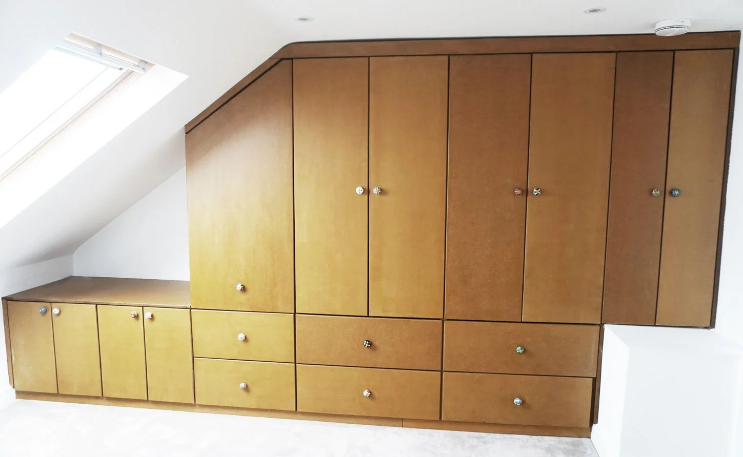 Wooden built-in cabinet with multiple doors and drawers under a sloped ceiling with a skylight window.