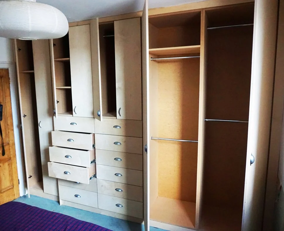Unfinished wooden closet with open cabinets, drawers, and empty hanging rods, next to a wooden door and purple bedding.