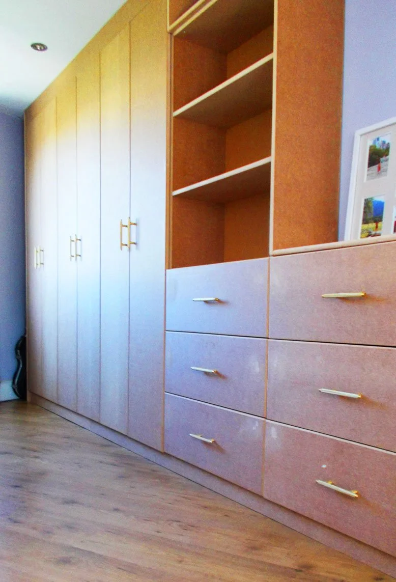 A built-in wooden wardrobe with closed doors and open shelves, located in a room with wooden flooring and a partially blue wall.