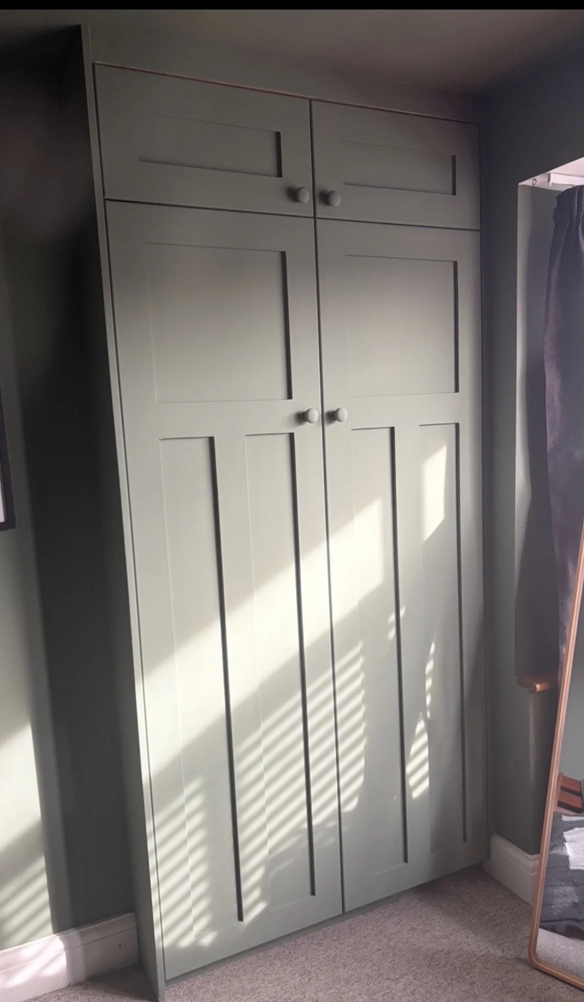A tall, light gray wardrobe with four doors and two smaller top drawers, situated next to a wall with sunlight casting shadows through window blinds. Part of a mirror and curtain are also visible.