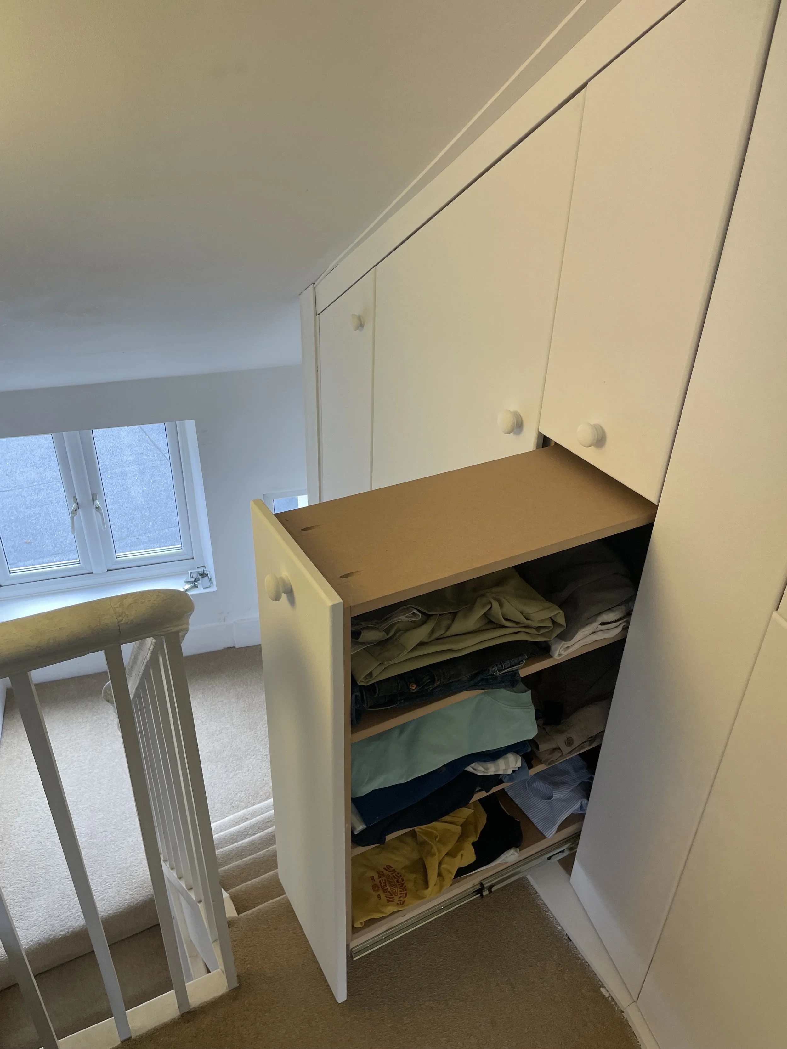 Open drawer of white built-in wardrobe with folded clothes inside, next to staircase railing and window.
