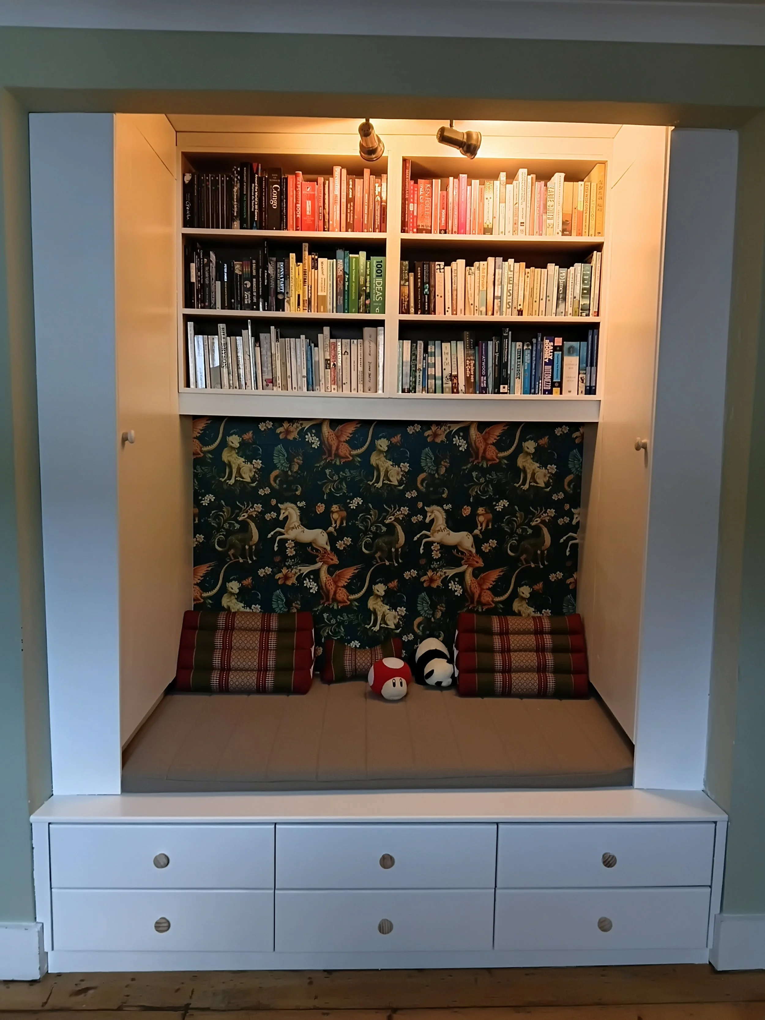 White built-in wardrobe with open door revealing a decorative room inside. The interior features a bookshelf filled with colorful books, a patterned fabric wallpaper with images of unicorns and dragons, and three small cushions with stripes and polka