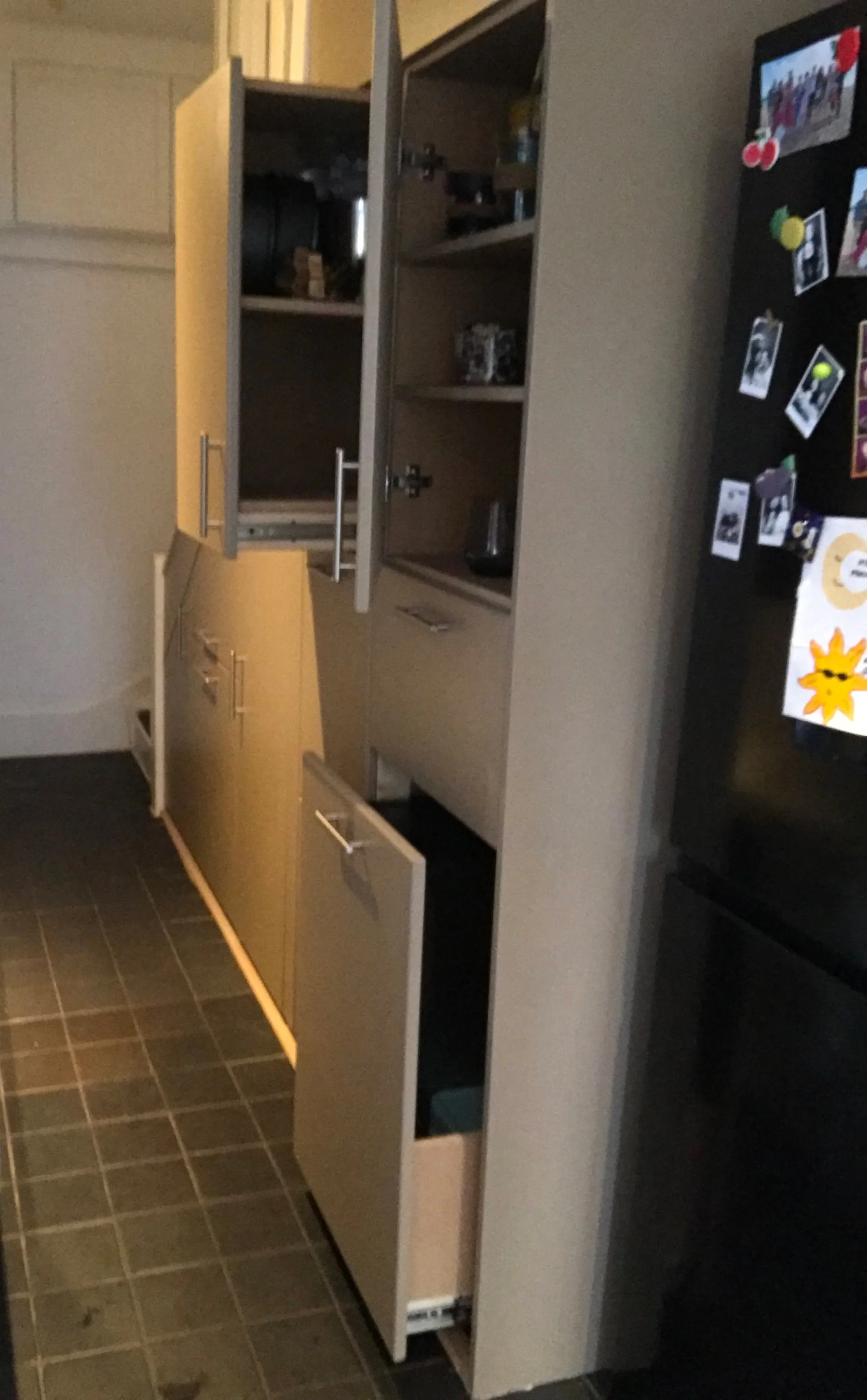 Open kitchen cabinet with shelves and a closed drawer below, located near a black refrigerator with magnets and photos.