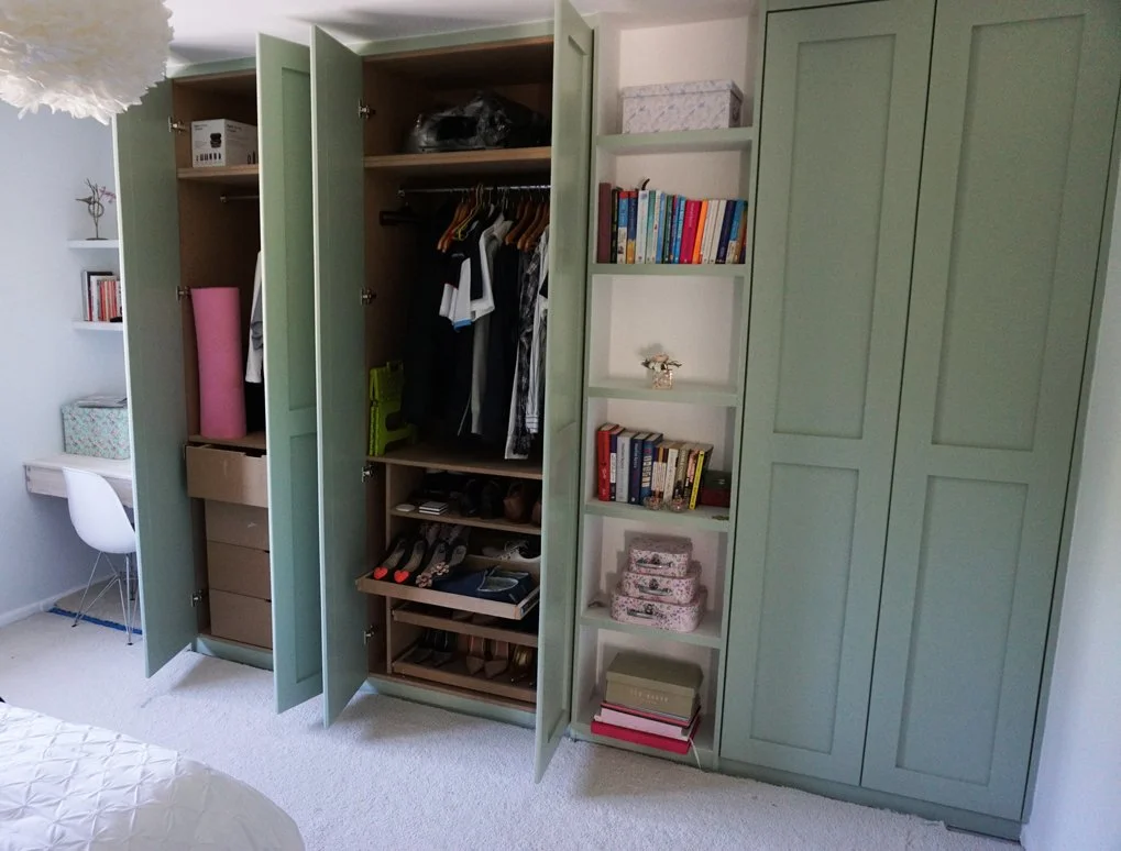 Open mint green wardrobe with clothes, shoes, and storage boxes inside, next to white bookshelf with books and decorative items, in a bedroom.