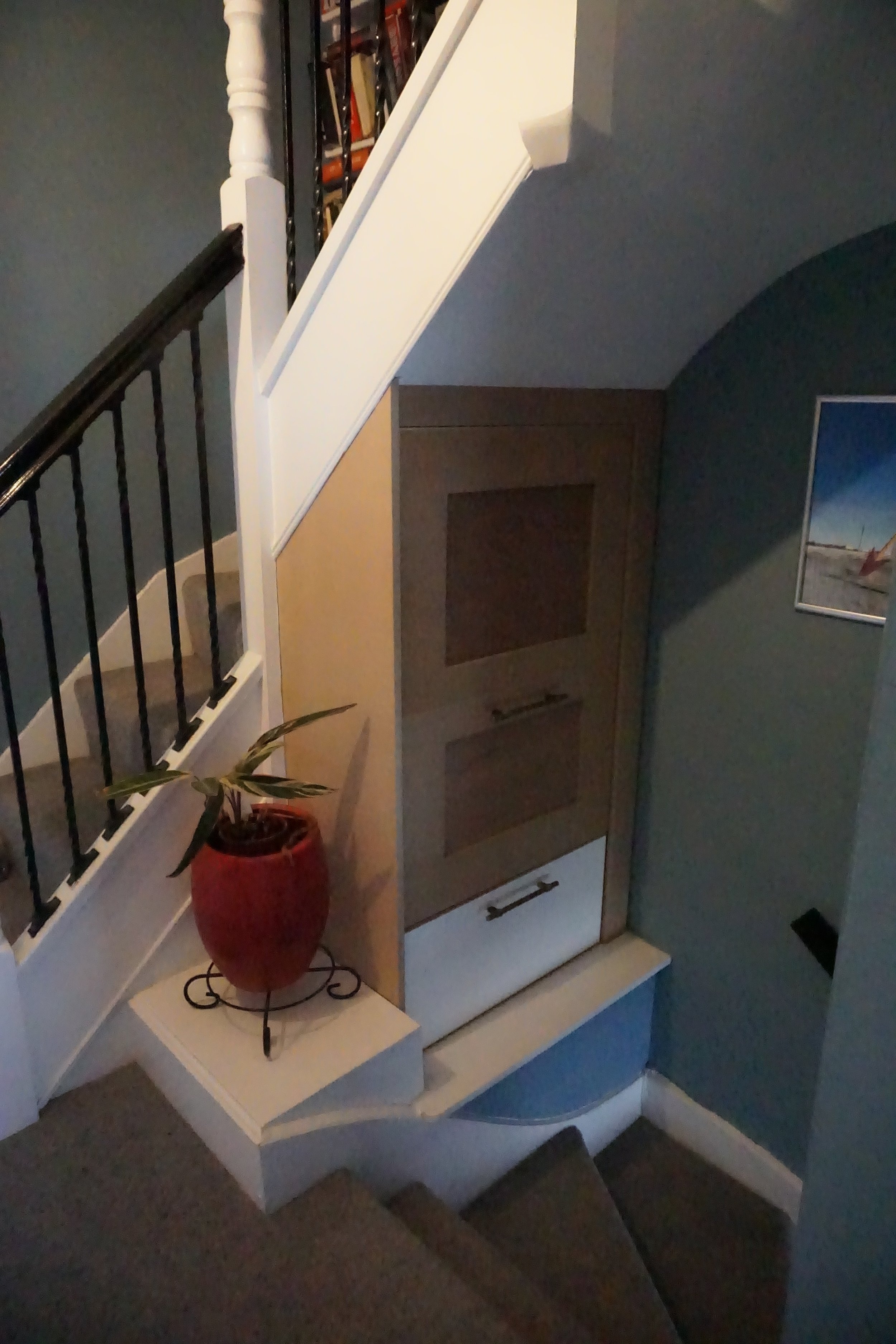 Staircase landing with a red potted plant on a white shelf, a built-in wooden cabinet, blue wall with framed photo, and carpeted stairs leading down.