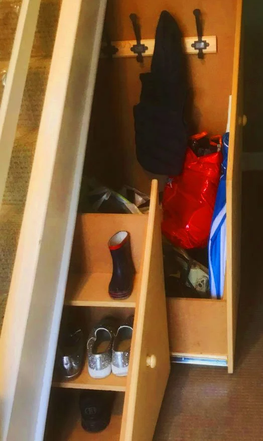 A small wooden entryway organizer with hooks, compartments, and shoes stored inside, including a black jacket hanging on the hooks, a red bag, a blue bag, a black rubber boot, and pairs of sneakers.