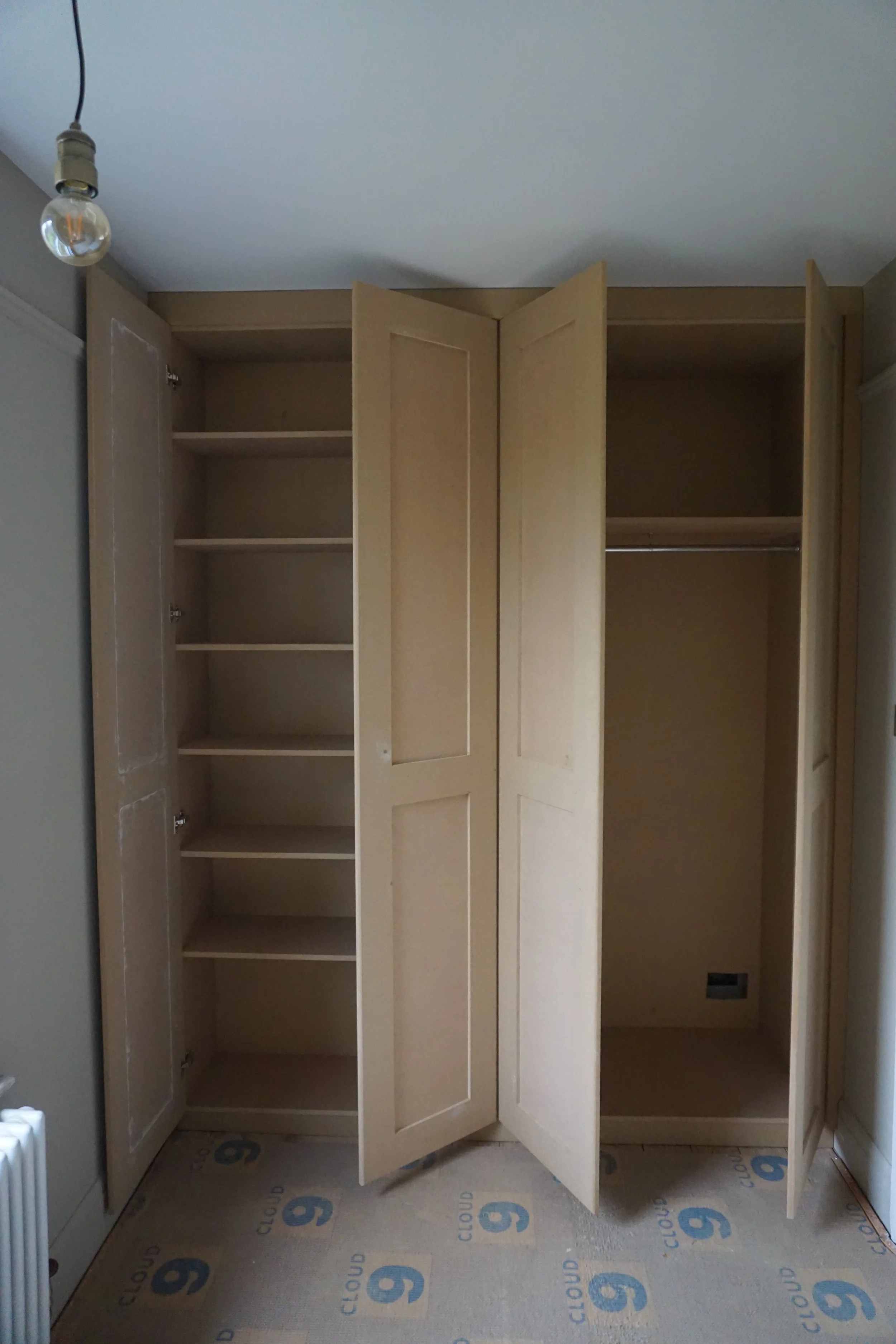 Empty built-in wardrobe with open doors, showing multiple shelves and a hanging rod inside. The wardrobe is beige and situated in a room with a grey wall and a ceiling light fixture.