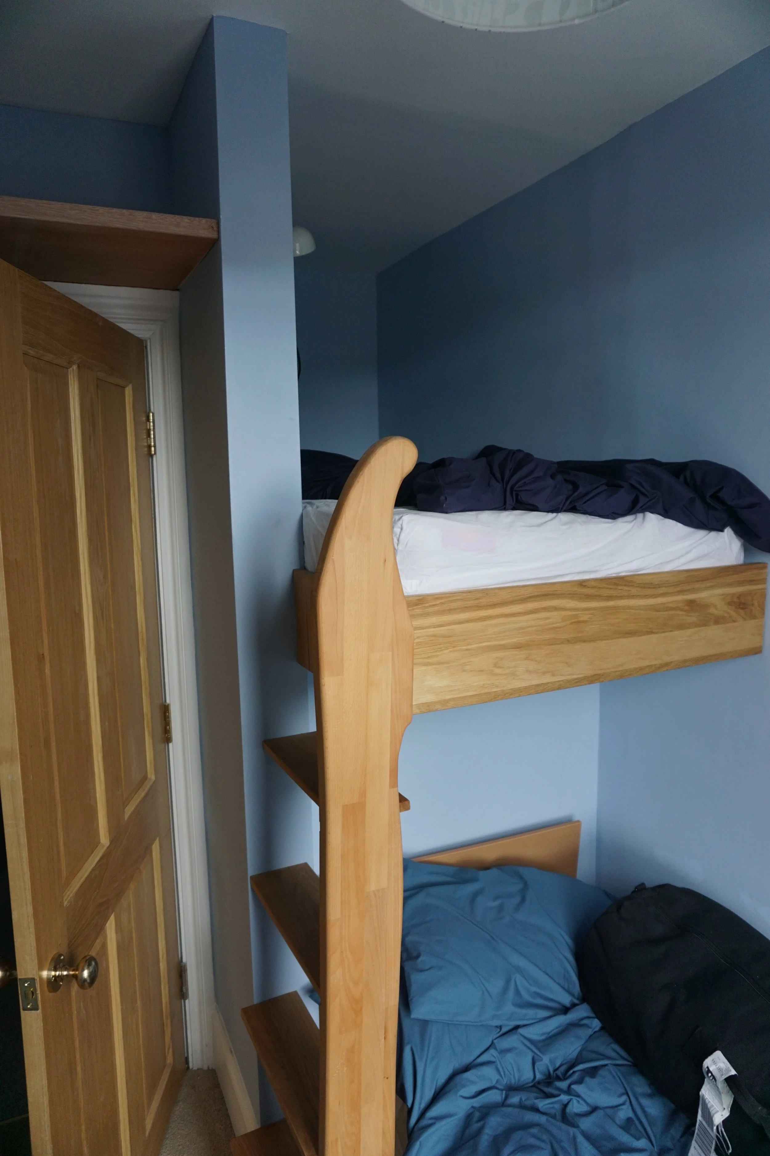 A wooden loft bed with a ladder in a blue-walled bedroom, with a mattress on top and a blue bedsheet and black bag on the bottom bed.