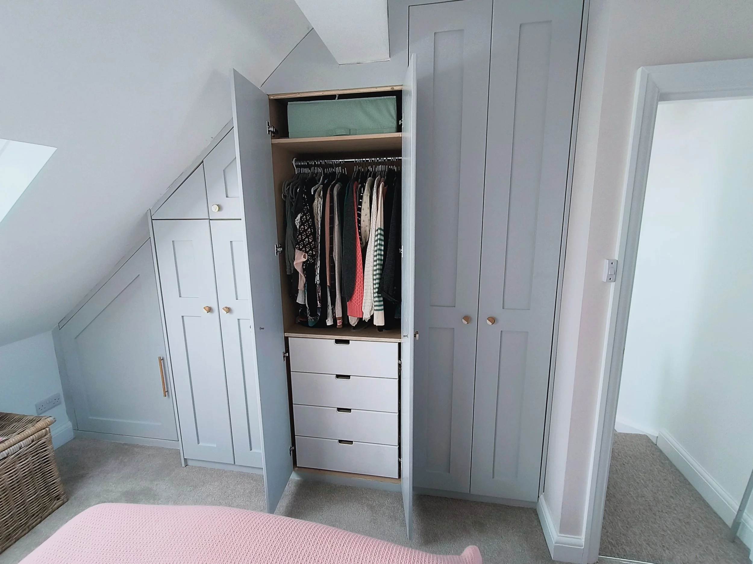 Open closet with clothing hanging inside, drawers below, and cabinets around it in a bedroom with sloped ceiling.