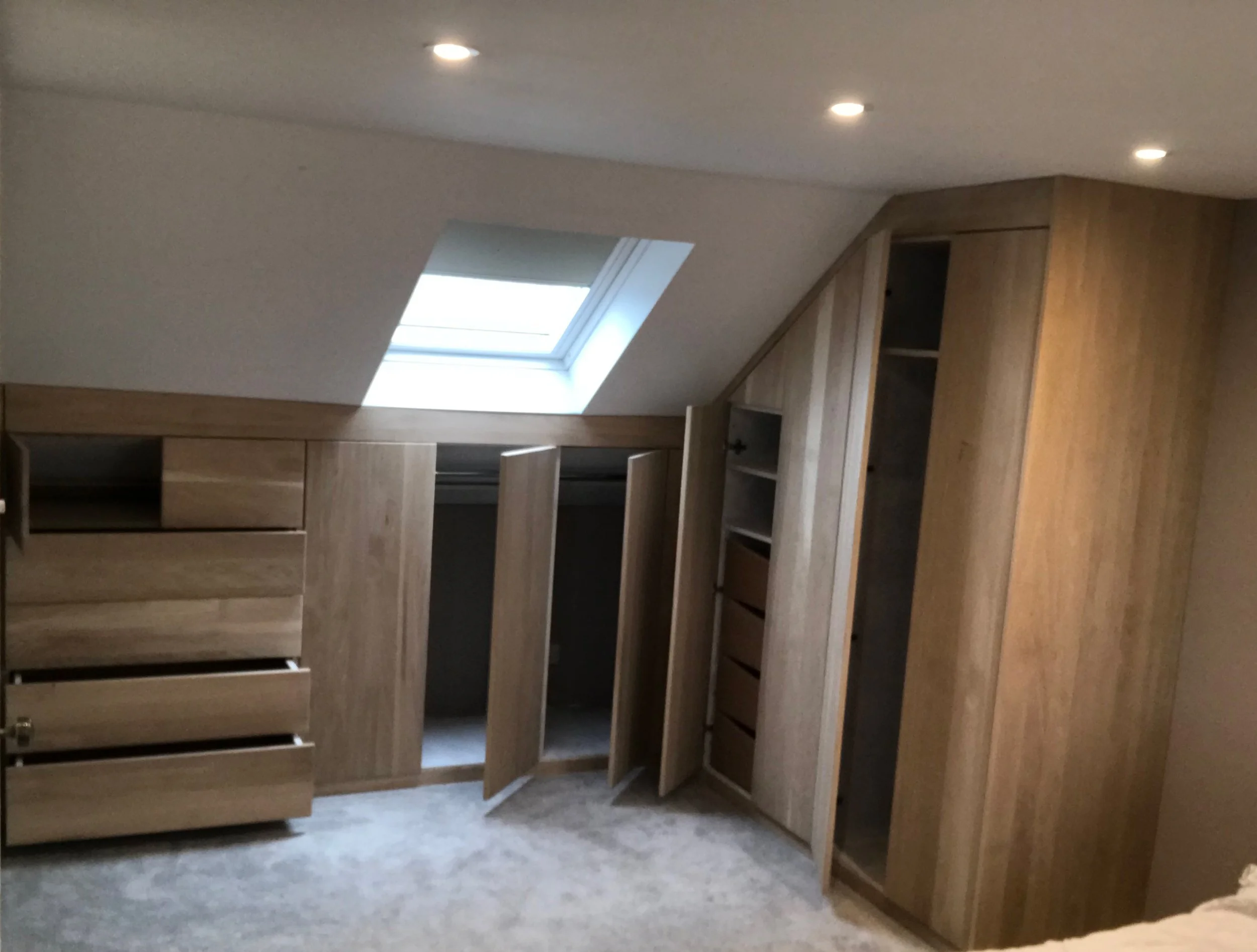 Wooden closet units with open drawers and cabinets under a sloped ceiling with a skylight window and ceiling lights.