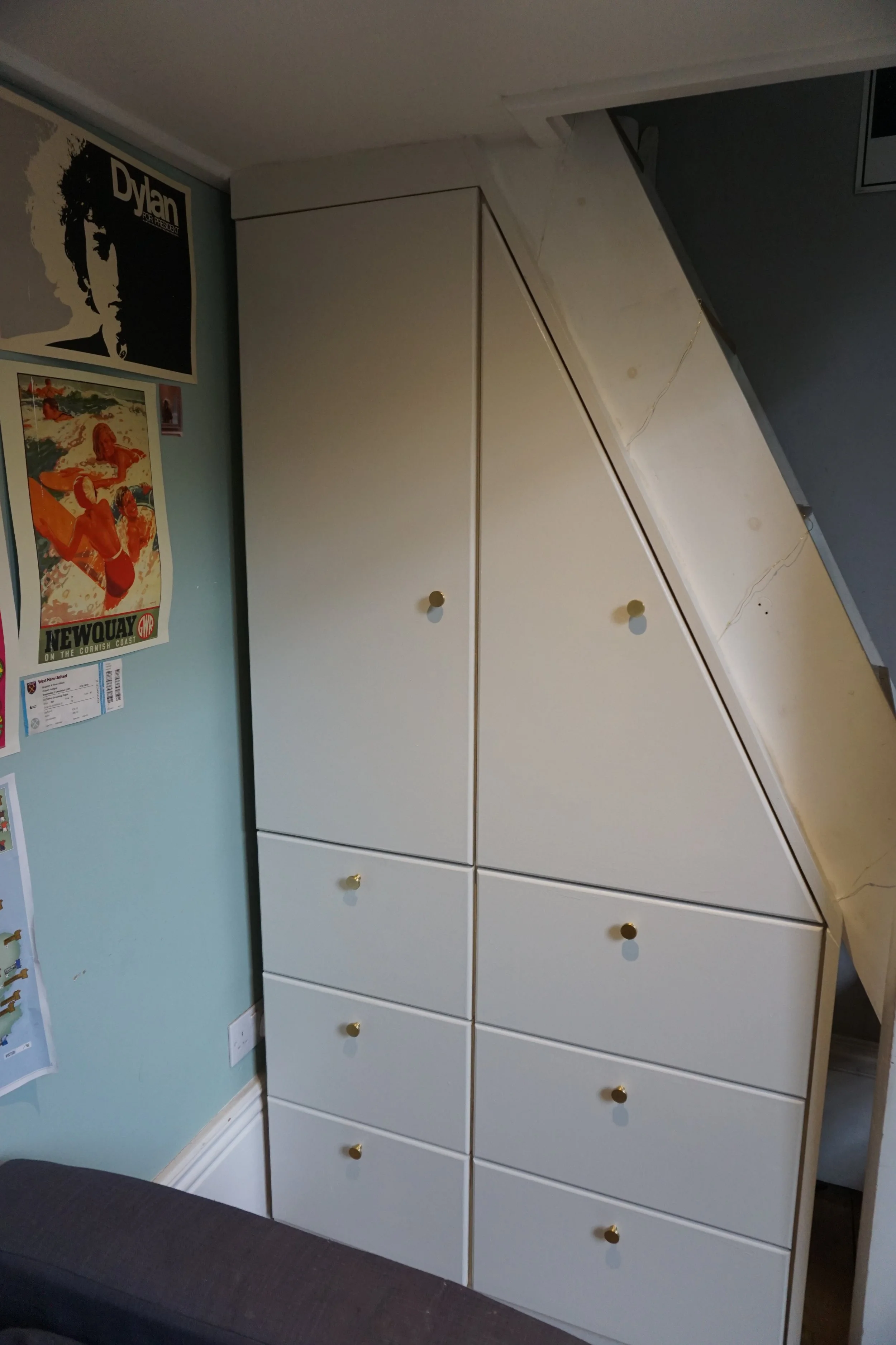 White wardrobe with multiple drawers and double doors, situated in a room with posters on the wall and a dark-colored sofa at the bottom left.