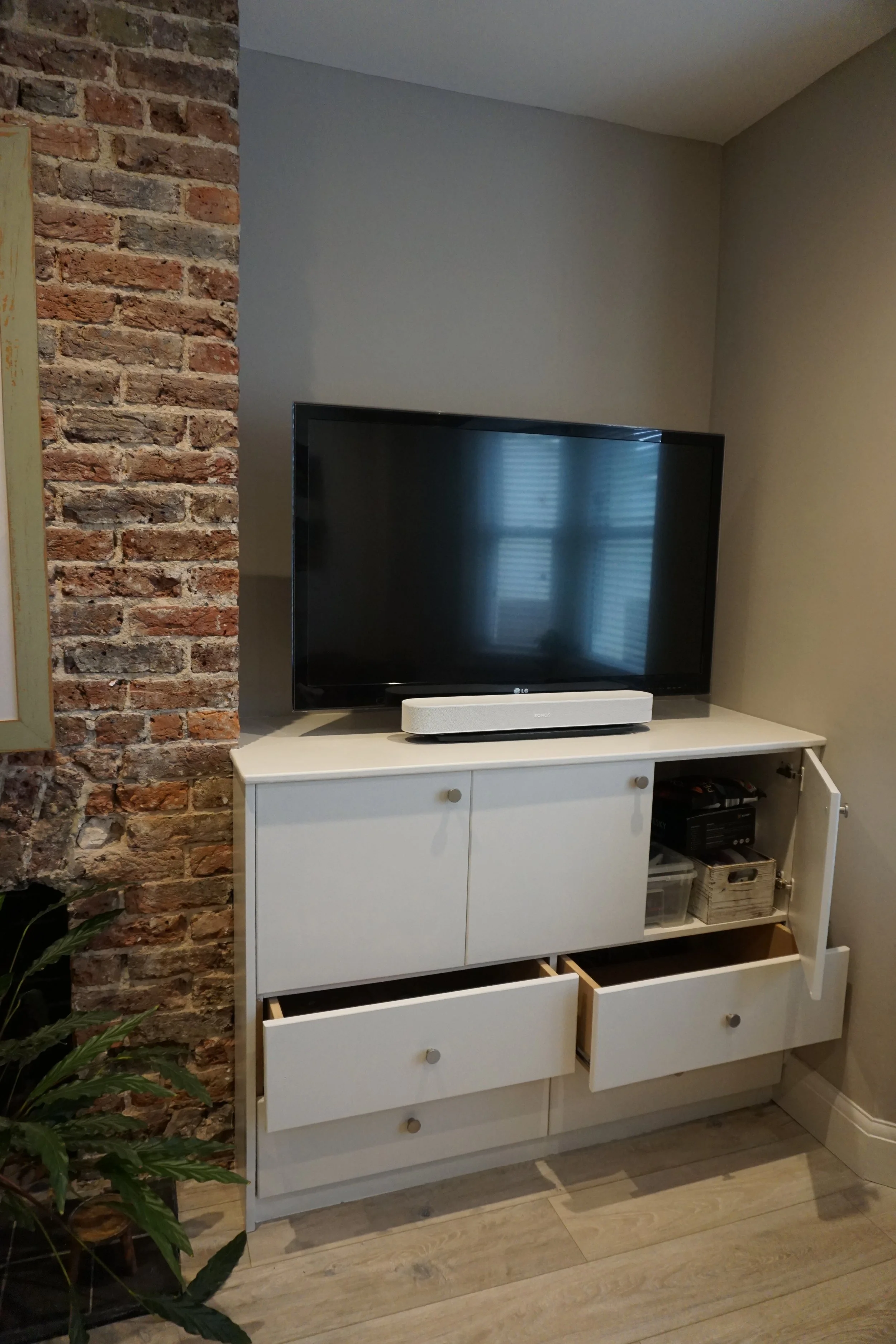 Flat-screen TV on a white cabinet with drawers, with a soundbar in front and a closed cabinet door to the right, against a wall with a brick accent on the left.
