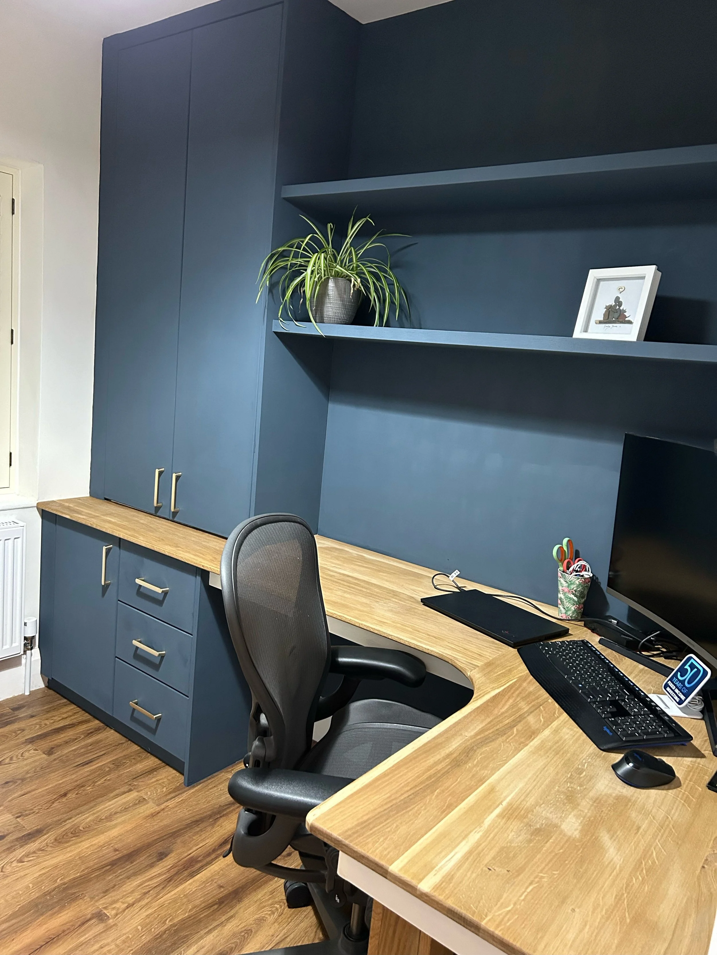 Home office with a wooden desk, blue cabinets, a black office chair, a computer monitor, a keyboard, a mouse, and a cup with scissors. There is a plant and a framed picture on the wall shelves.