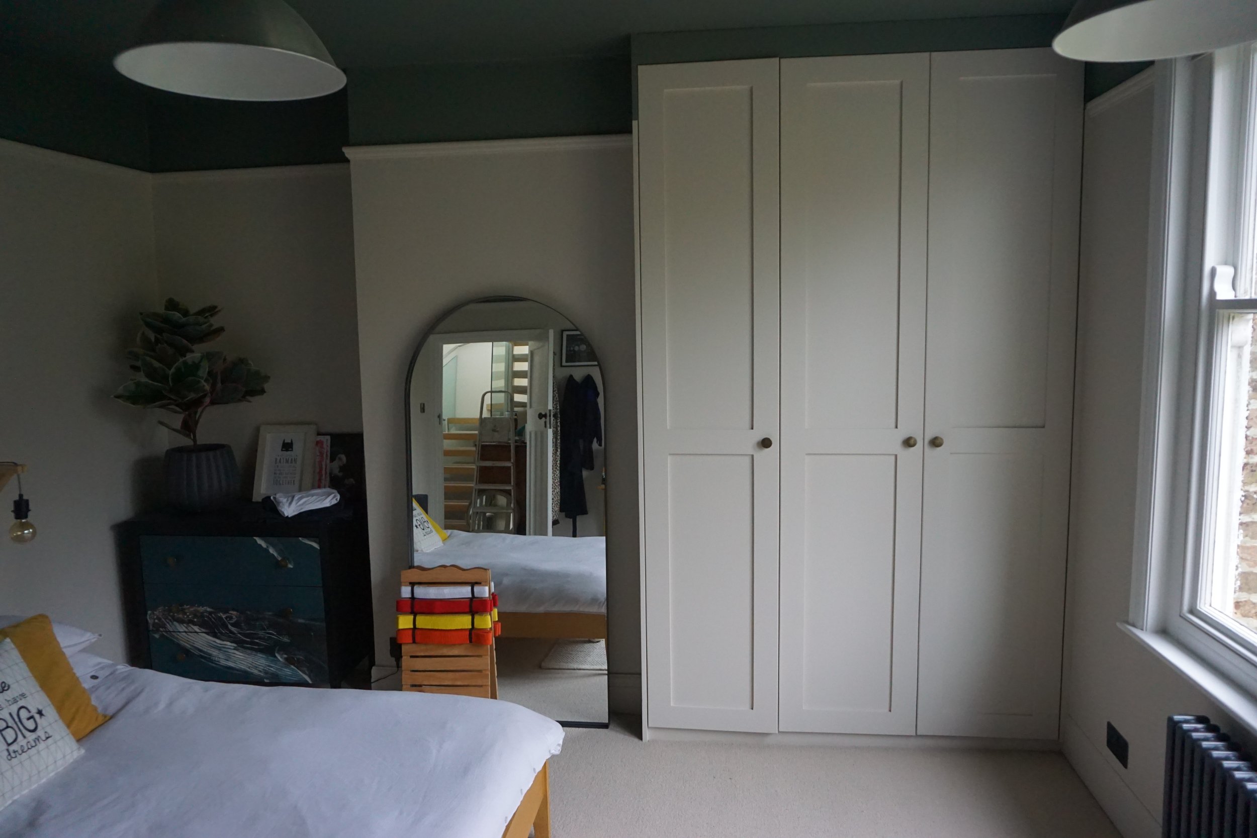 A bedroom with white bedding, a blue dresser, a large potted plant, a round mirror, white built-in wardrobes, and a window with natural light.