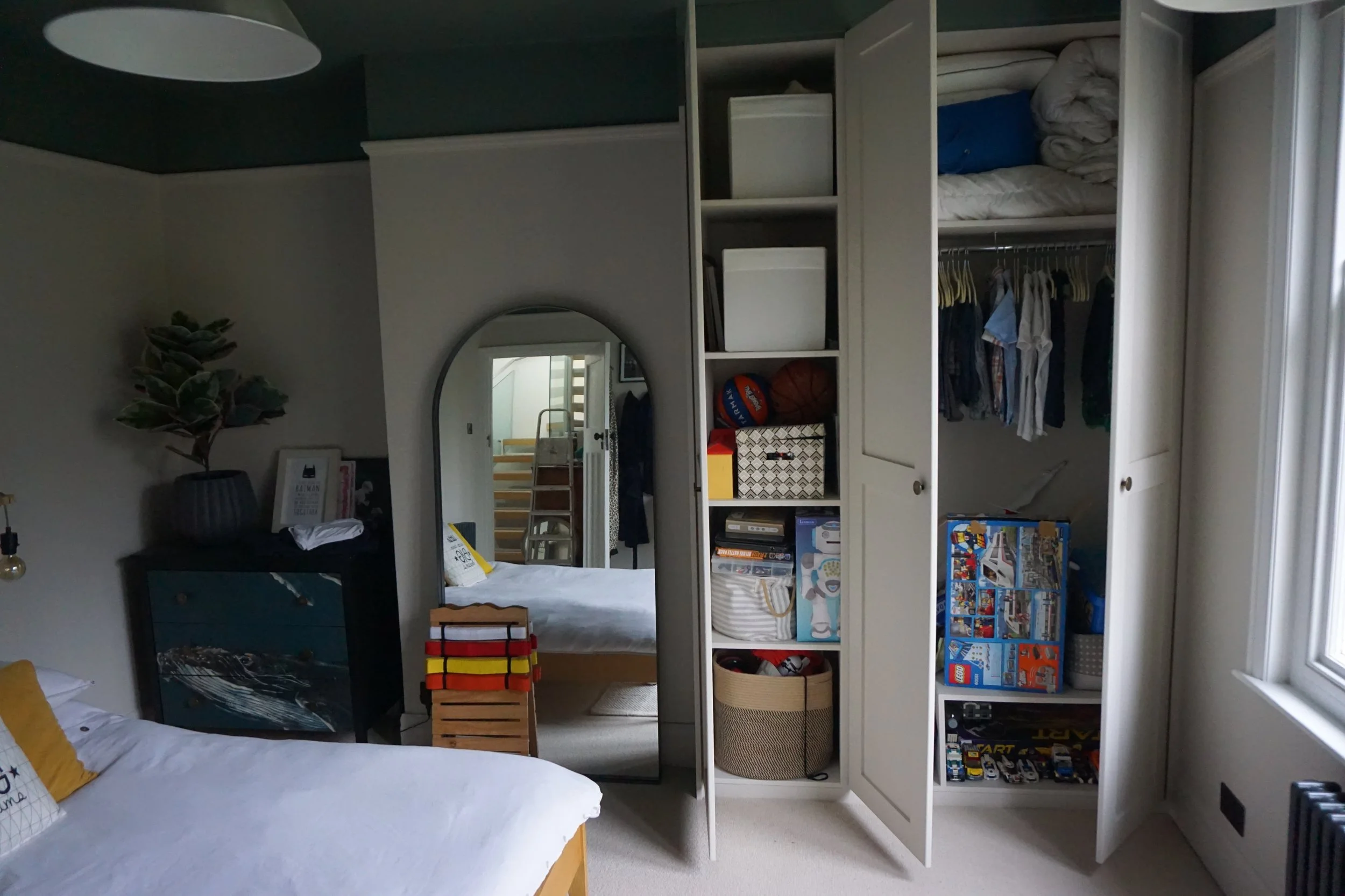 Bedroom with open closet, showing folded bed linens, hanging clothes, toys, and storage boxes. Part of a bed with a yellow pillow, and a mirror reflecting a staircase.