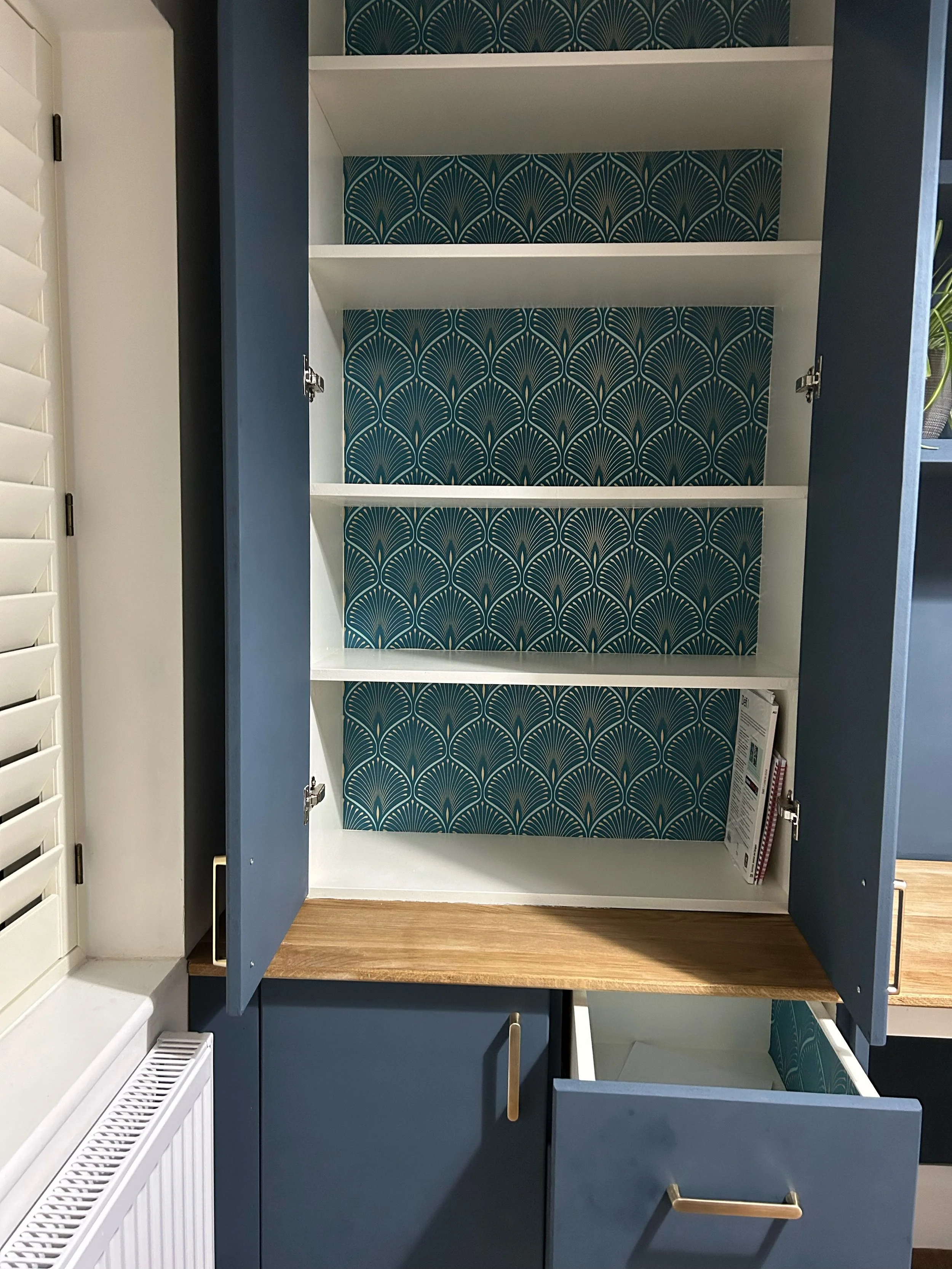 Open kitchen cabinet with white interior, blue exterior, and gold handles; interior shelves with blue and gold patterned wallpaper, containing some magazines.