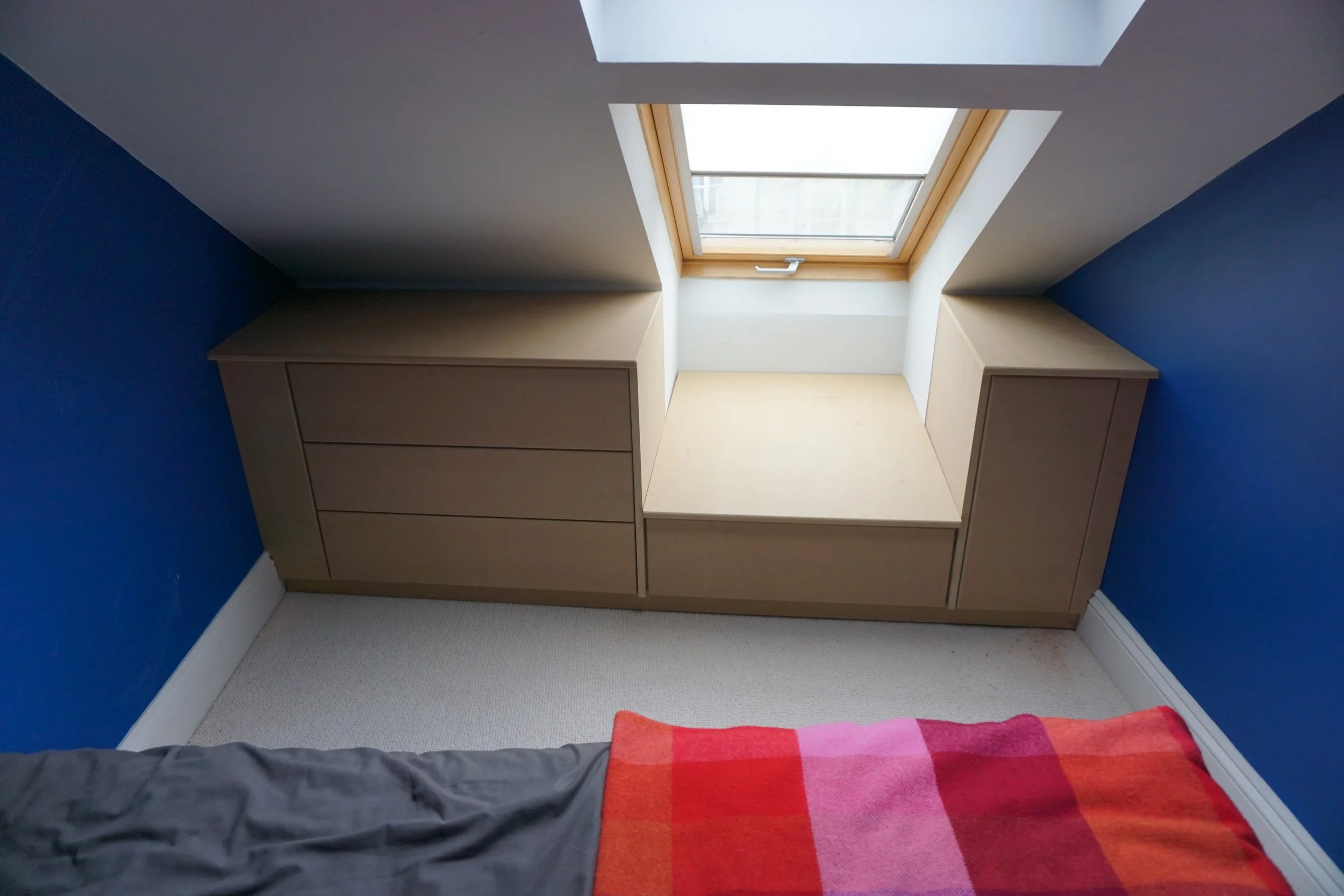 View of a bedroom corner with a built-in wooden cabinet under a skylight window, blue walls, white ceiling, beige carpet, and part of a bed with red and pink striped blanket.