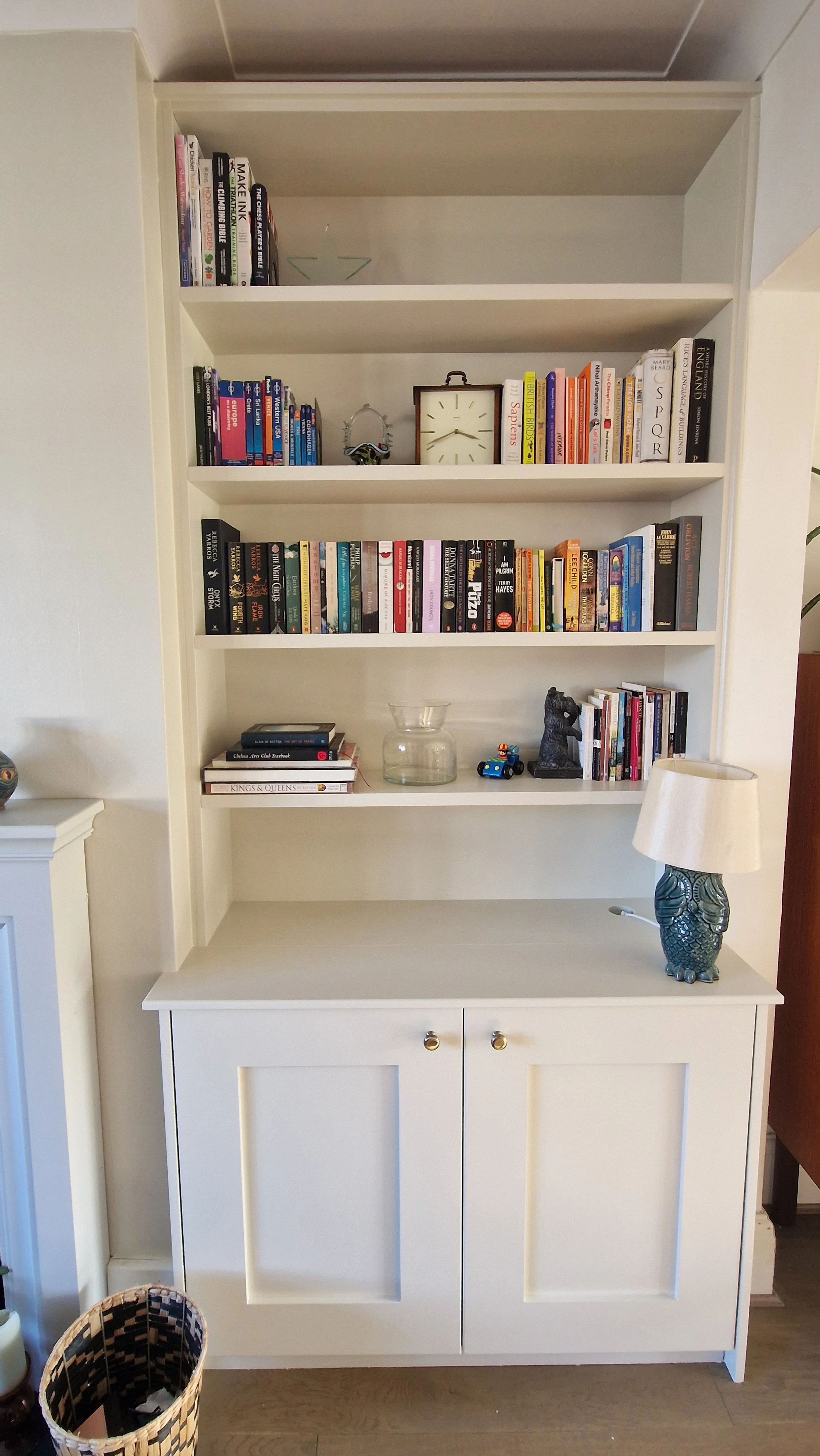 A white bookshelf with three shelves filled with books, decorative items, a glass vase, a small toy car, and a black carved figure. There is a white cabinet with two doors below the shelves and a table lamp with a blue owl-shaped base to the right. A
