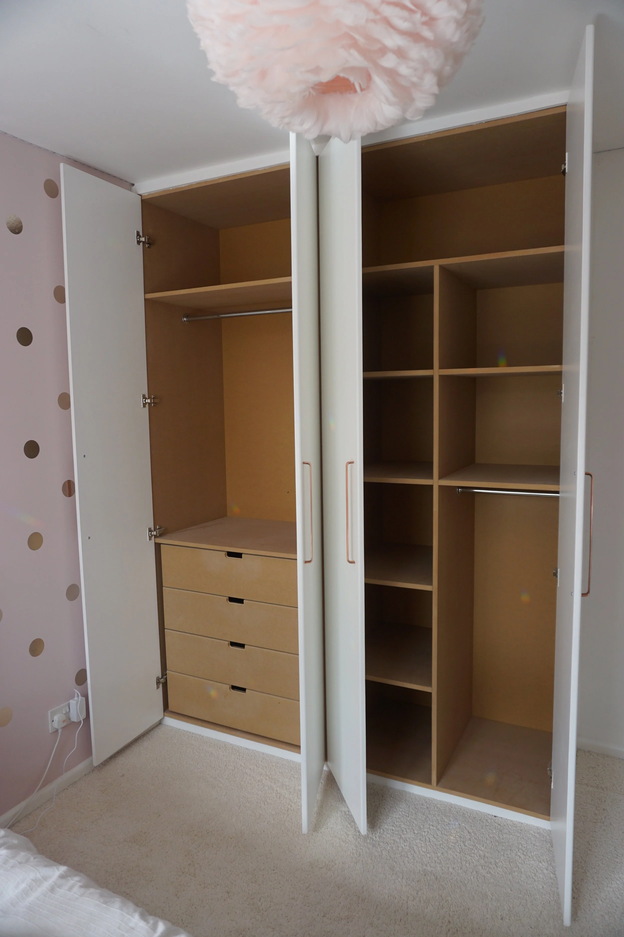 Open wardrobe closet with drawers on the left and multiple shelves and hanging rods inside, located in a bedroom with polka dot wallpaper and pink ceiling fixture.
