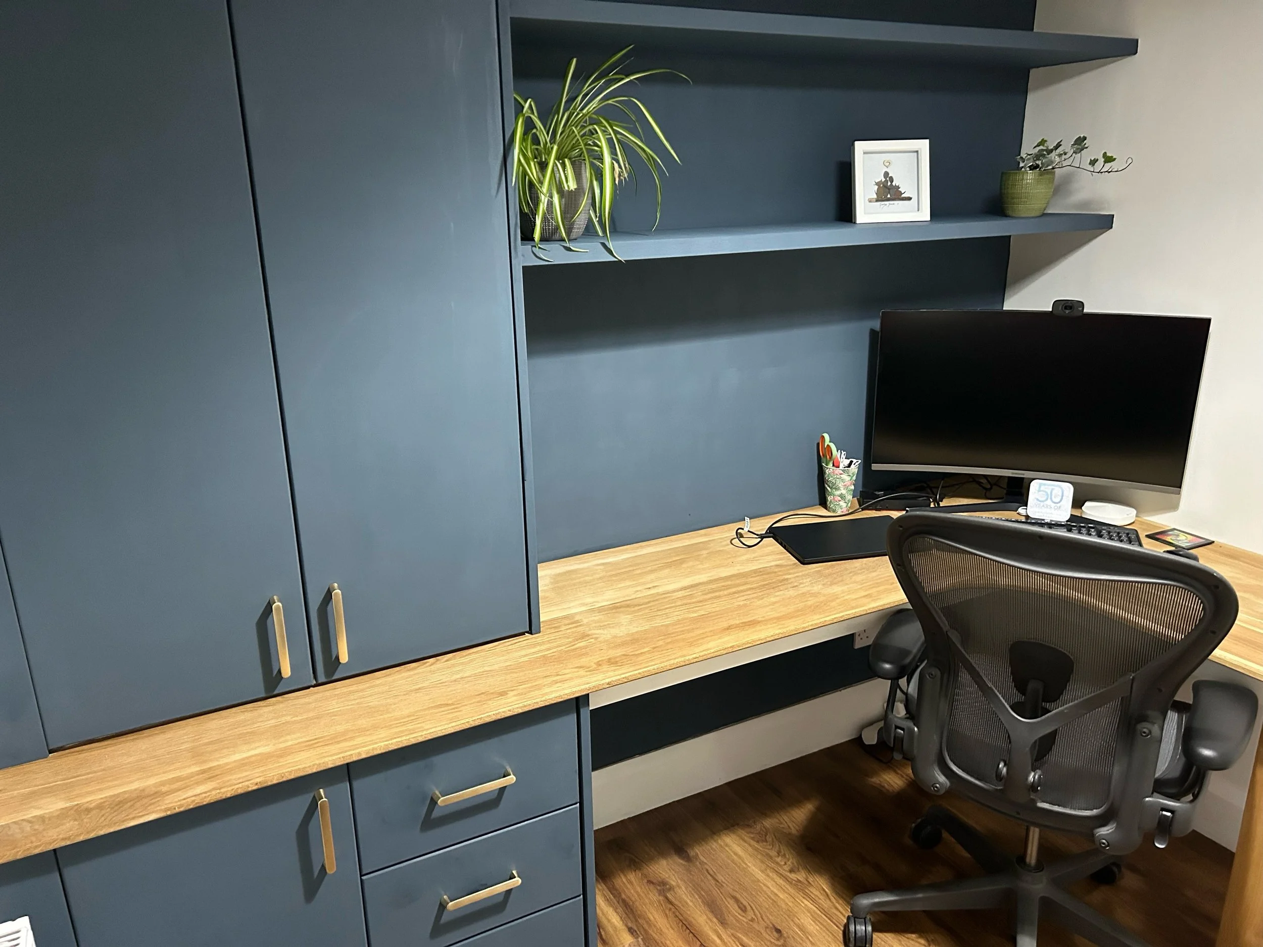 Home office with a wooden desk, black office chair, computer monitor, and built-in cabinetry with shelves and drawers painted in blue-green. There are two small potted plants on the shelves and a small picture frame.