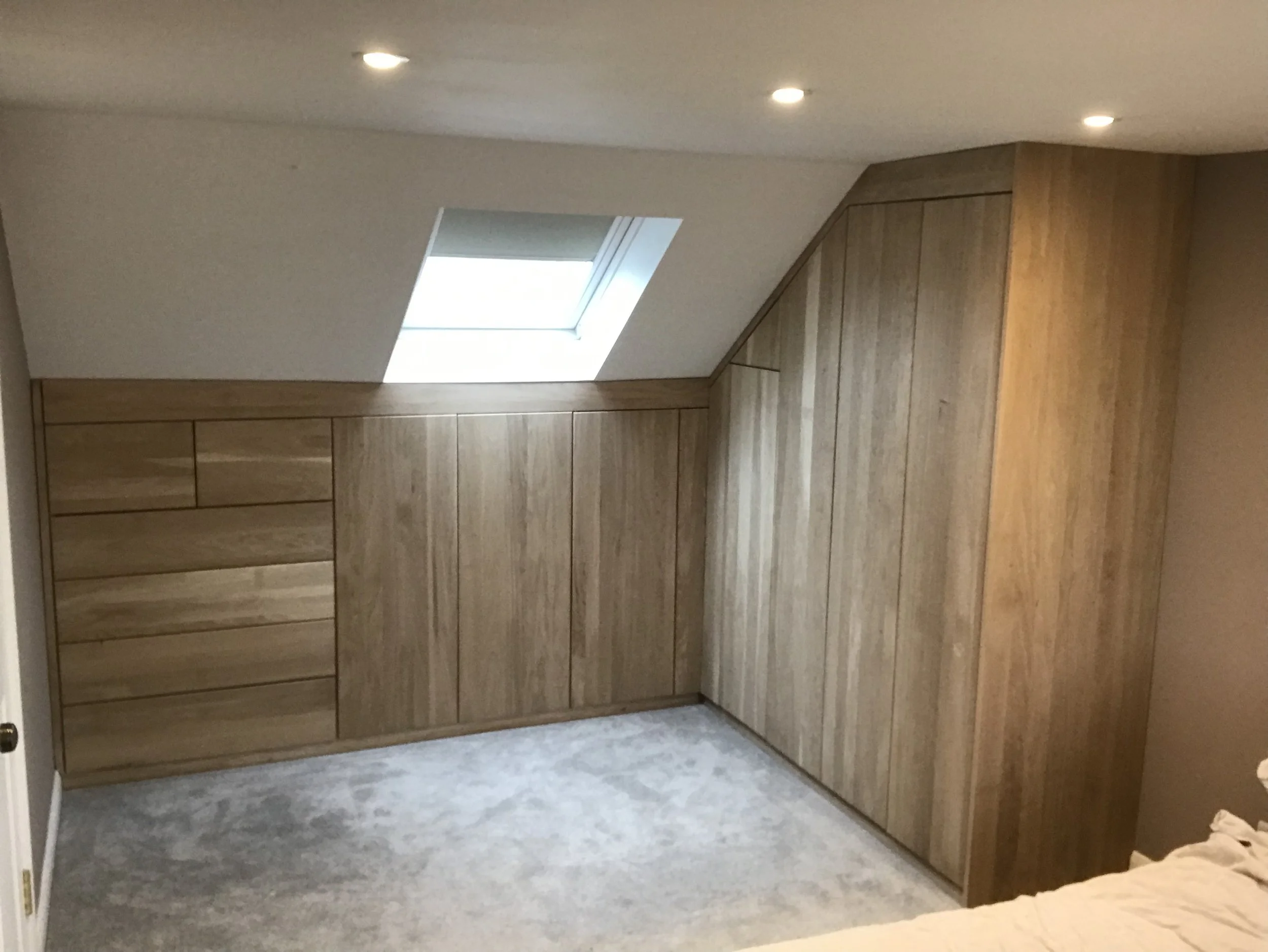 A room with wooden closet cabinets and a skylight window on the sloped ceiling. The floor is carpeted, and part of a bed is visible in the lower right corner.