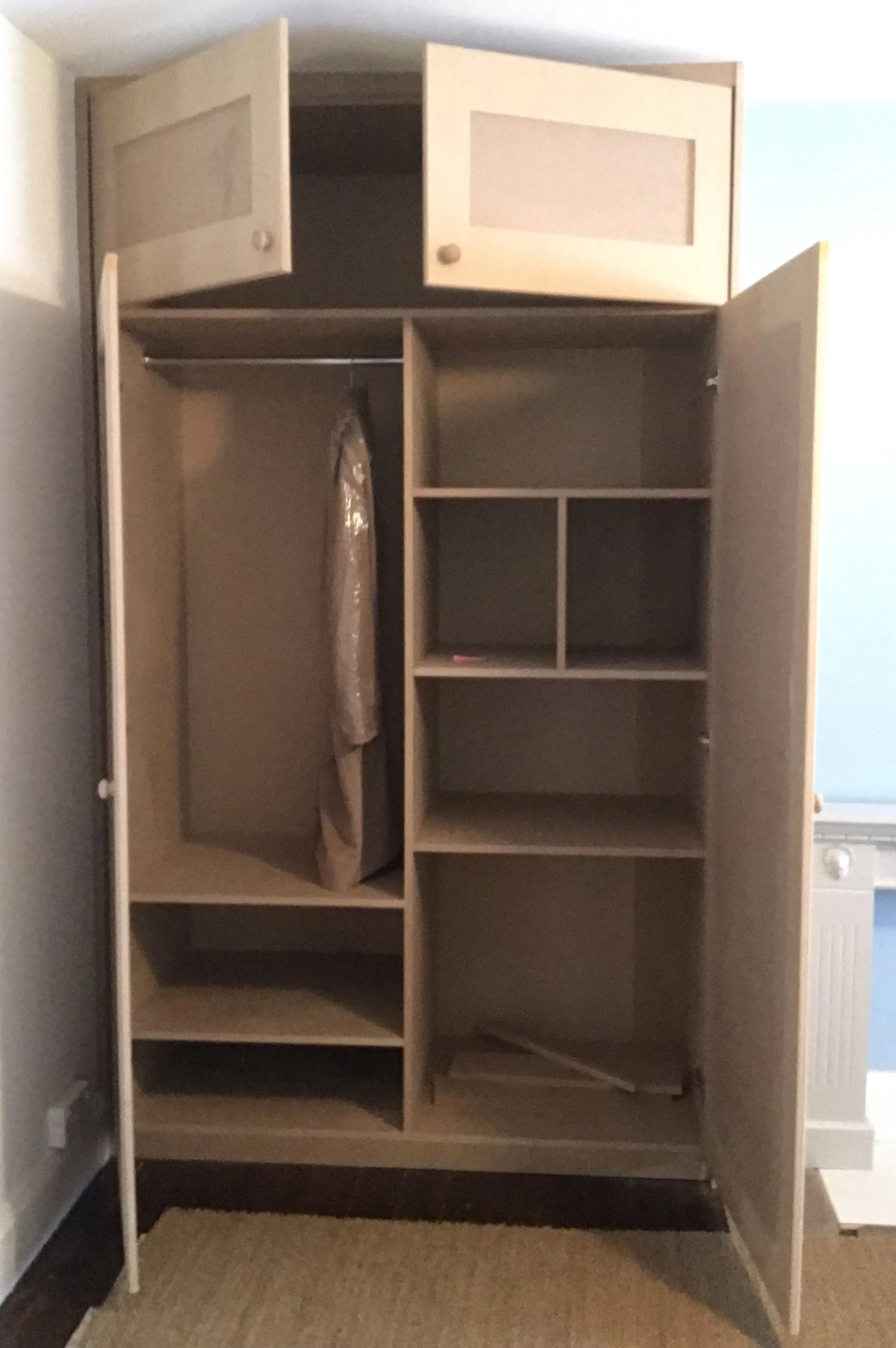 Empty wooden wardrobe with open doors, showing shelves and a hanging rod inside, located in a room with light-colored walls and a beige rug.