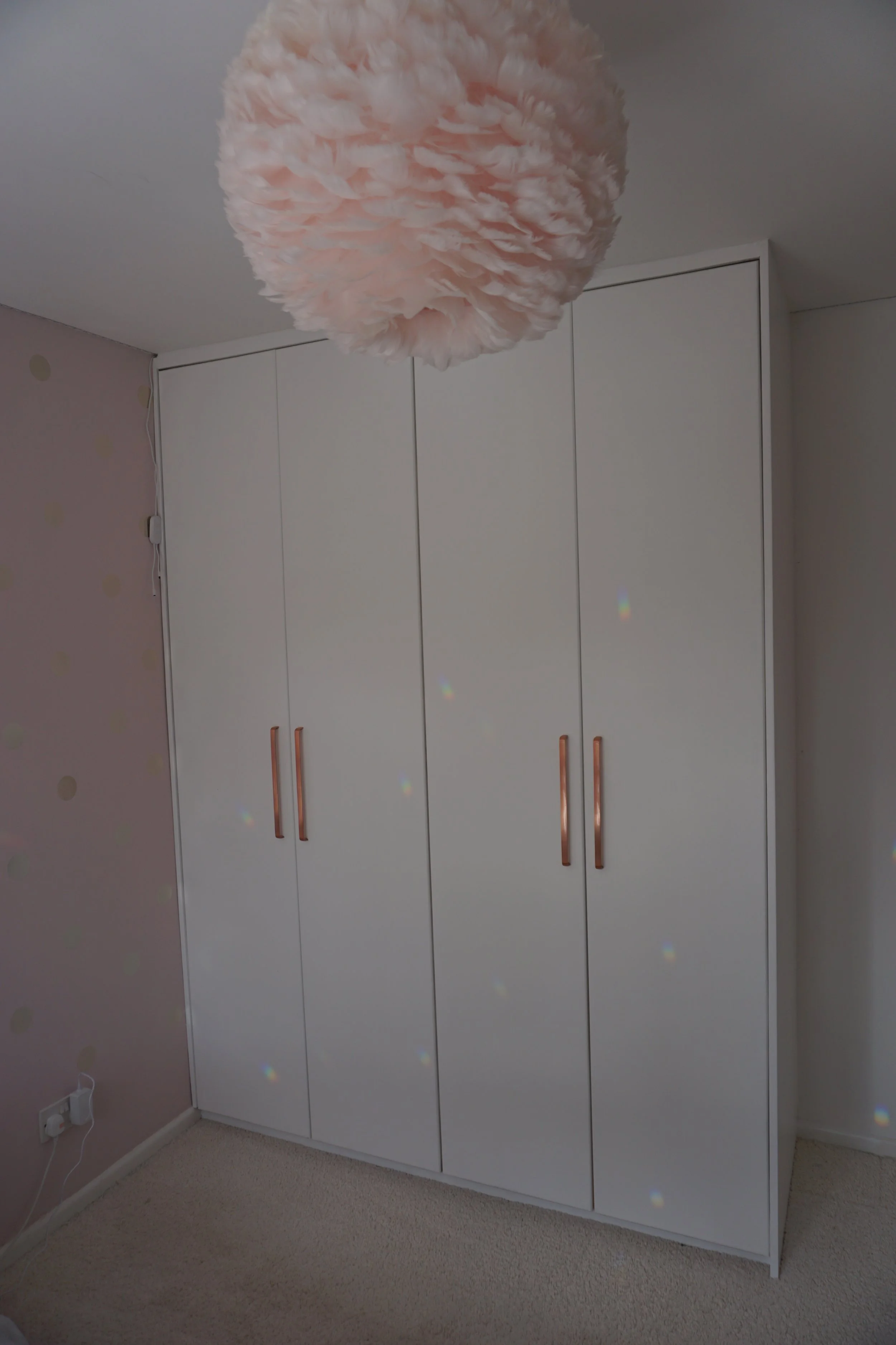 White wardrobe with four doors and rose gold handles in a room with pink polka-dotted wall, beige carpet, and a pink fluffy ceiling light.