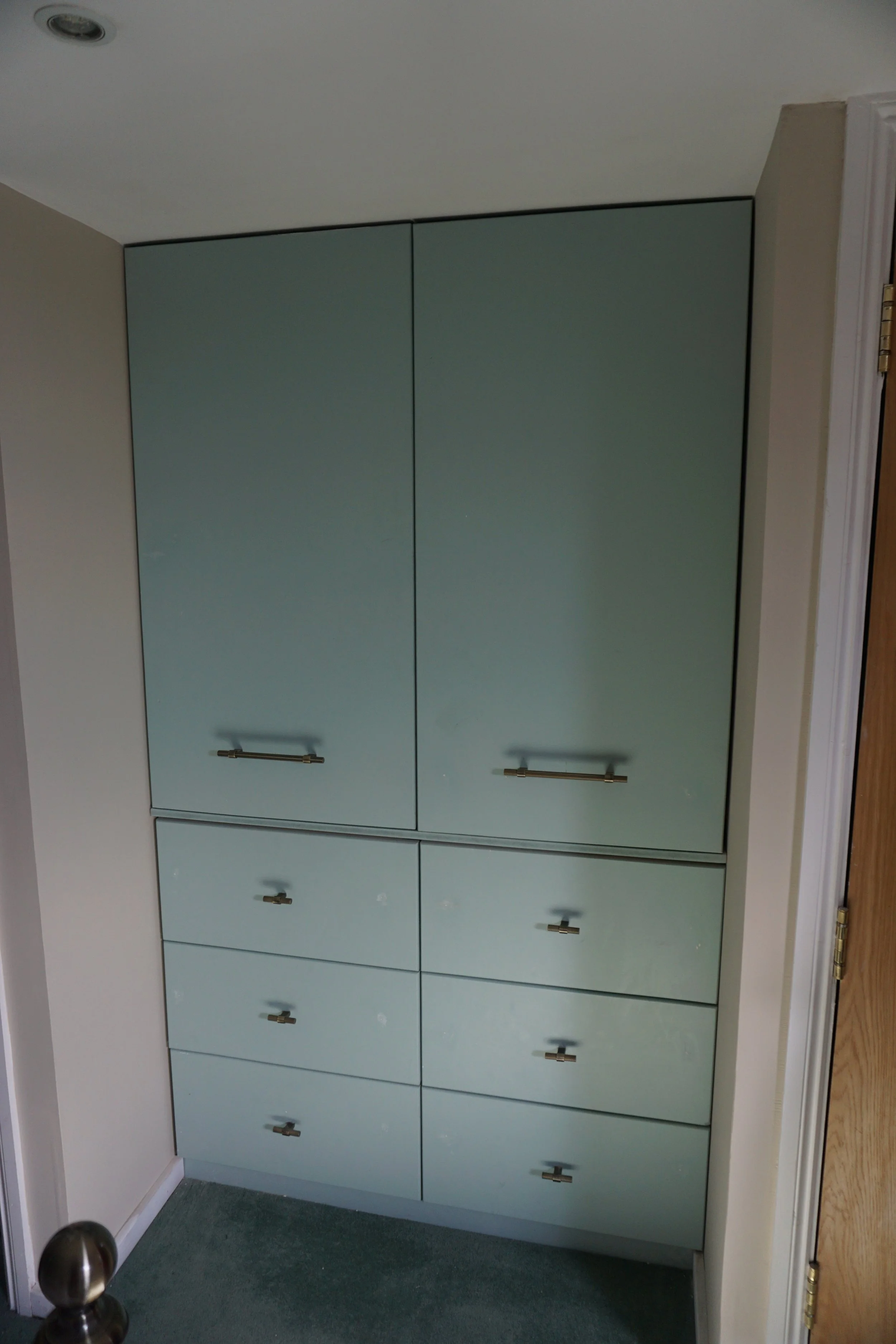 A built-in cabinet with six drawers at the bottom and two large doors above, positioned in a room corner. The cabinet has a light green color and metallic handles.