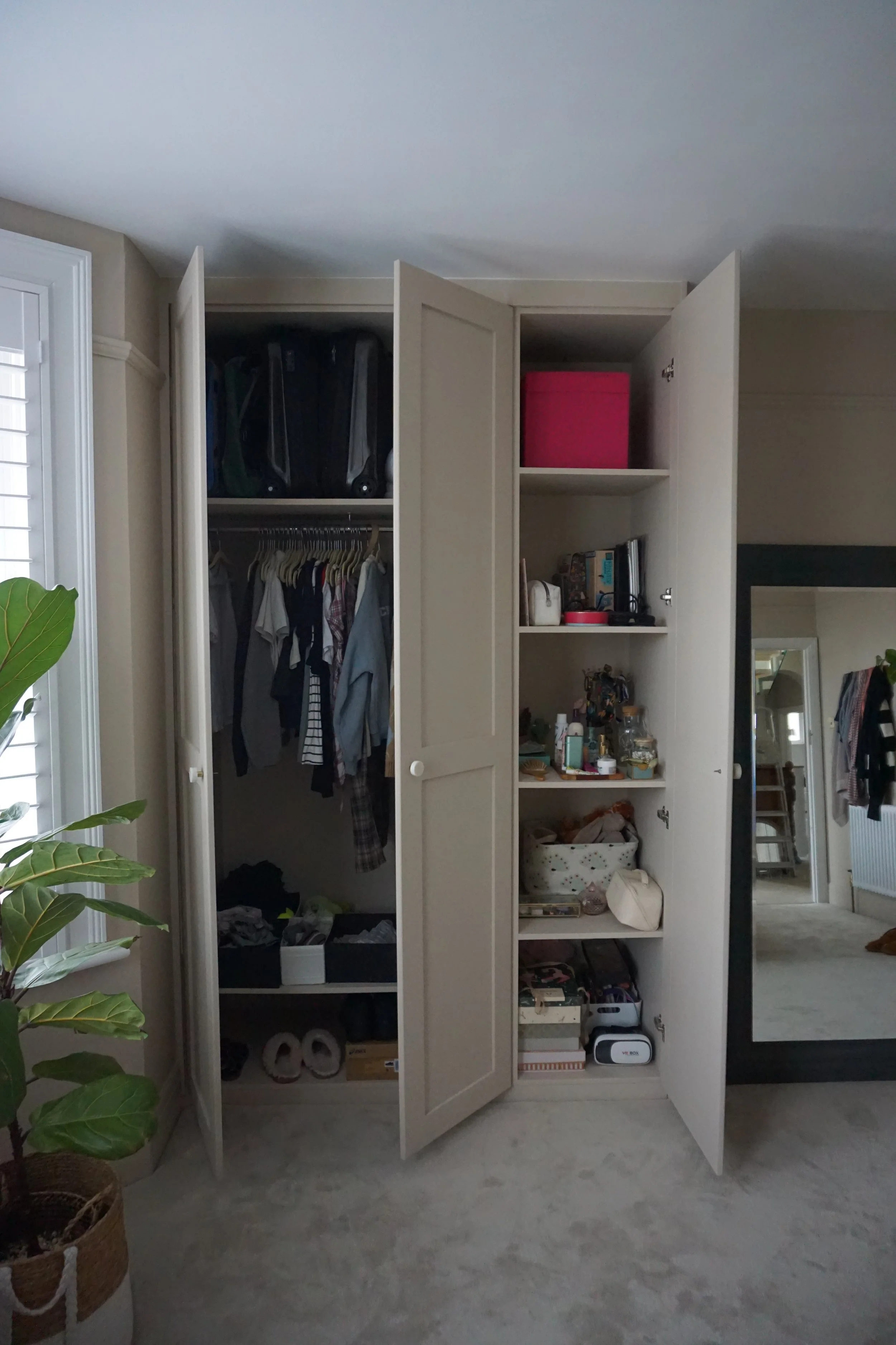 Open closet with clothes hanging, shelves with books, jars, and miscellaneous items, and a large mirror reflecting part of a room.